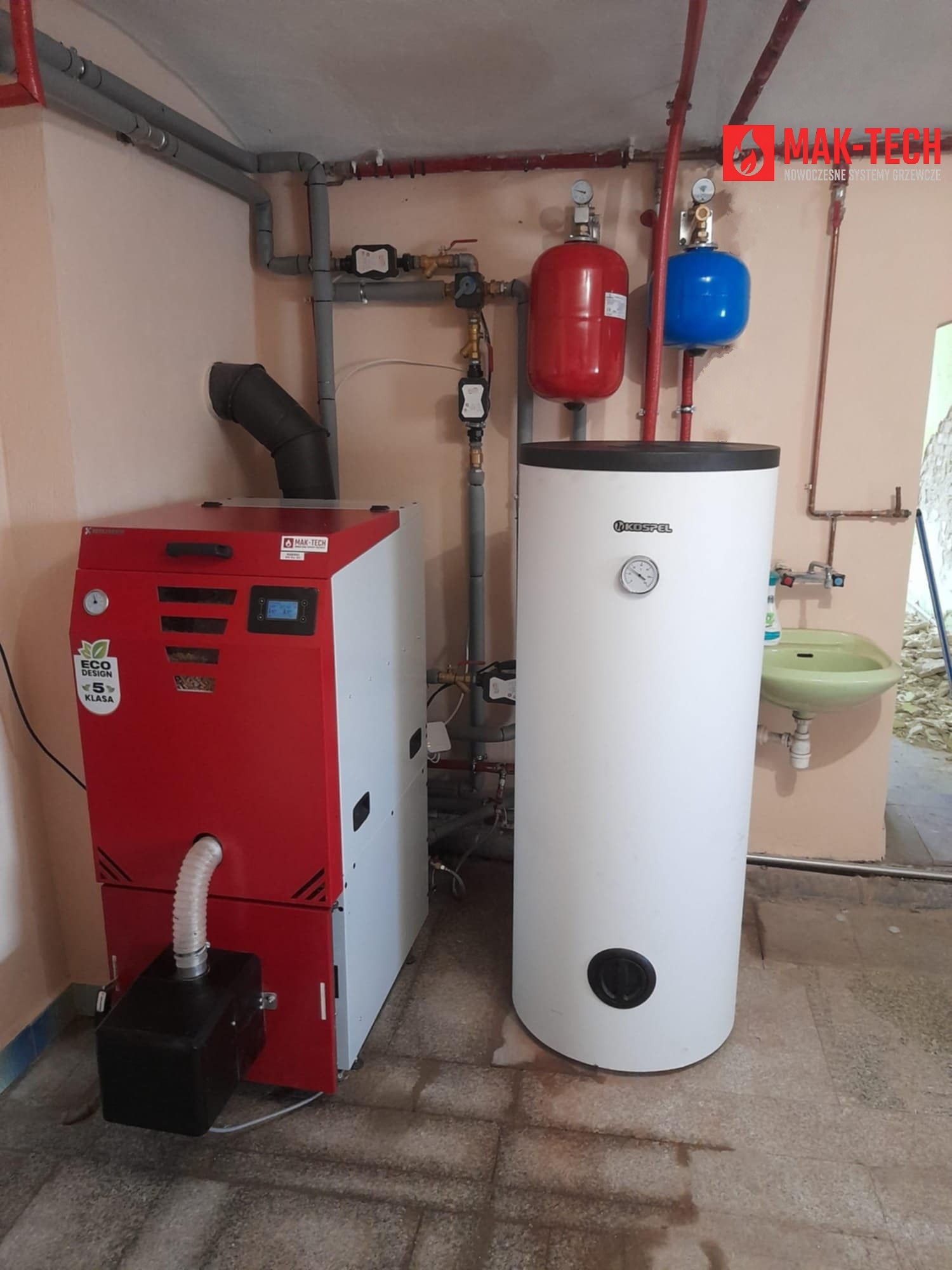 Czerwono-biały piec na paliwo stałe Eco Design klasy 5 z podajnikiem, obok biały bojler Kospel. Widoczne rury instalacji grzewczej i elementy hydrauliczne.