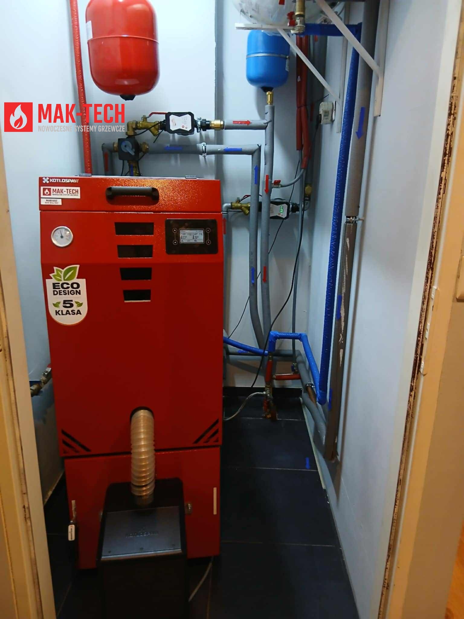 Czerwony piec na paliwo stałe marki MAK-TECH, z widocznymi elementami instalacji grzewczej, w małym pomieszczeniu kotłowni. Widoczne rury i zbiorniki wyrównawcze.