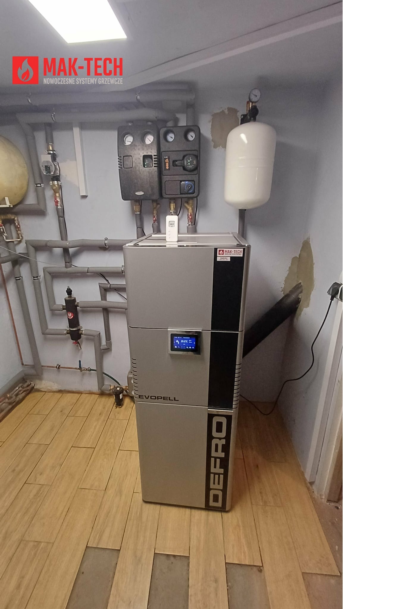 Szary piec na pellet marki Defro Evopell w kotłowni z widocznymi rurami instalacji grzewczej i zbiornikiem wyrównawczym. Na ścianie sterowniki i manometry.