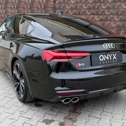 GHOST AUTODETAILING BIAŁYSTOK - Błyszczący, czarny Audi S5 po aplikacji powłoki ochronnej, widok z tyłu, prezentacja detali karoserii i felg na tle brązowej ściany.