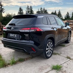 GHOST AUTODETAILING BIAŁYSTOK - Błyszczący, czarny samochód Toyota Corolla Cross Hybrid AWD po aplikacji powłoki ochronnej Onyx Coating, widok z tyłu na tle lasu i składowanego drewna.
