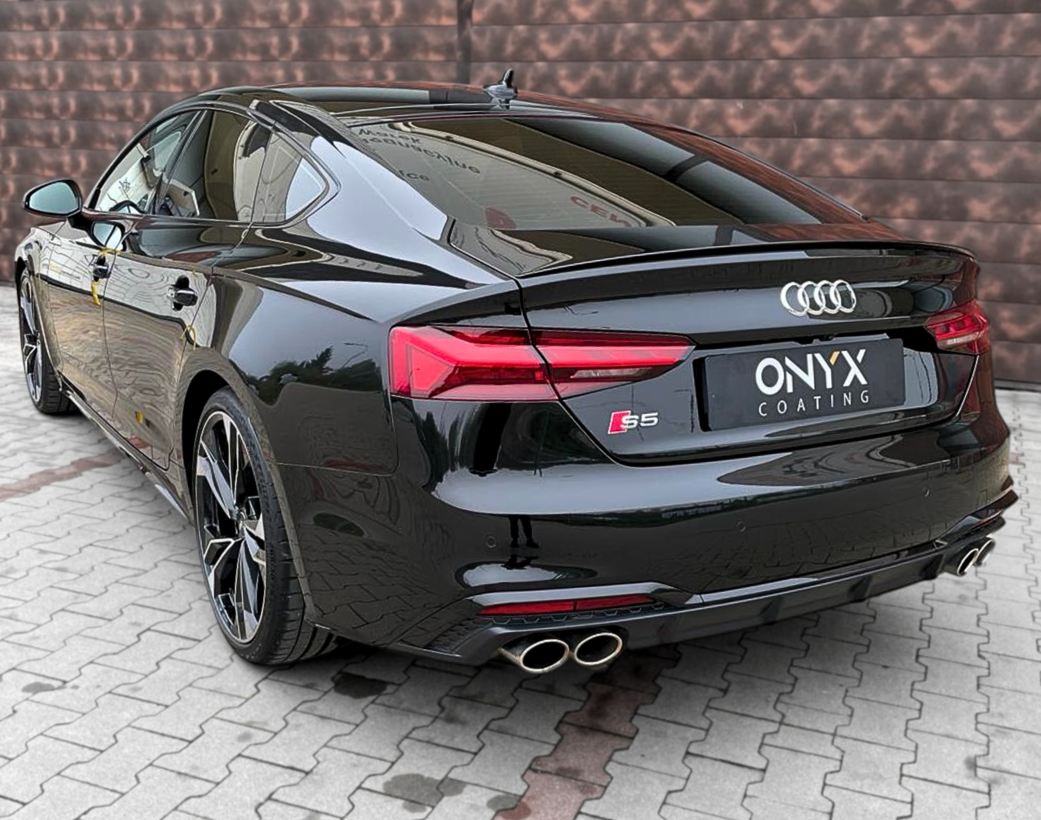 Błyszczący, czarny Audi S5 po aplikacji powłoki ochronnej, widok z tyłu, prezentacja detali karoserii i felg na tle brązowej ściany.
