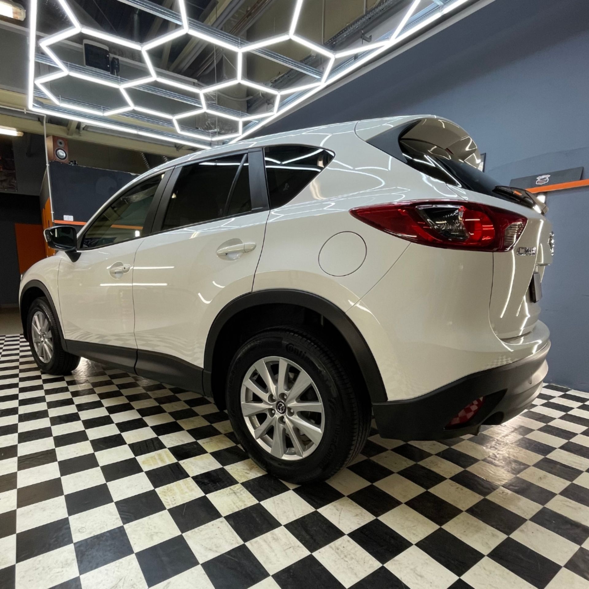 Biały samochód Mazda CX-5 po detailingu, stojący w garażu z czarno-białą szachownicą na podłodze i nowoczesnym oświetleniem LED w kształcie plastra miodu na suficie.