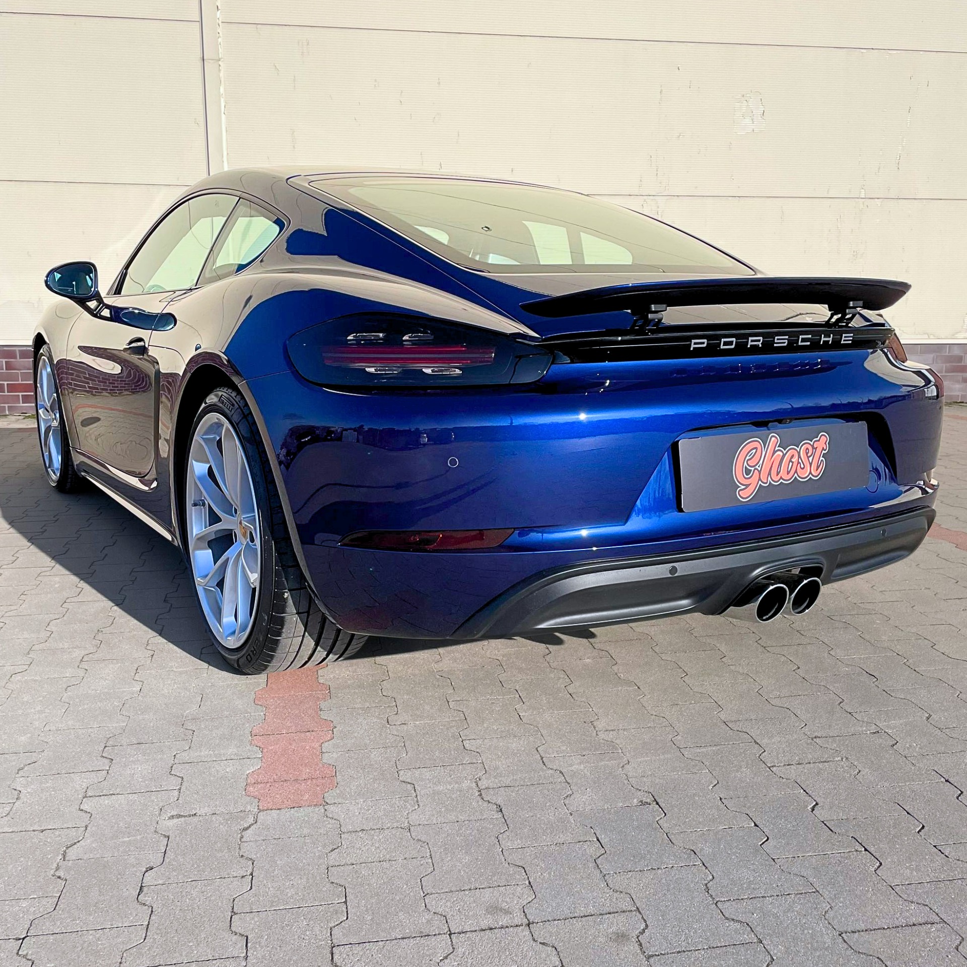 Błyszczący, granatowy Porsche Cayman z zamontowanym spojlerem, sfotografowany w Horodnianach z tyłu na tle jasnej ściany. Na tylnej szybie widoczna naklejka z nazwą firmy 'Ghost'.