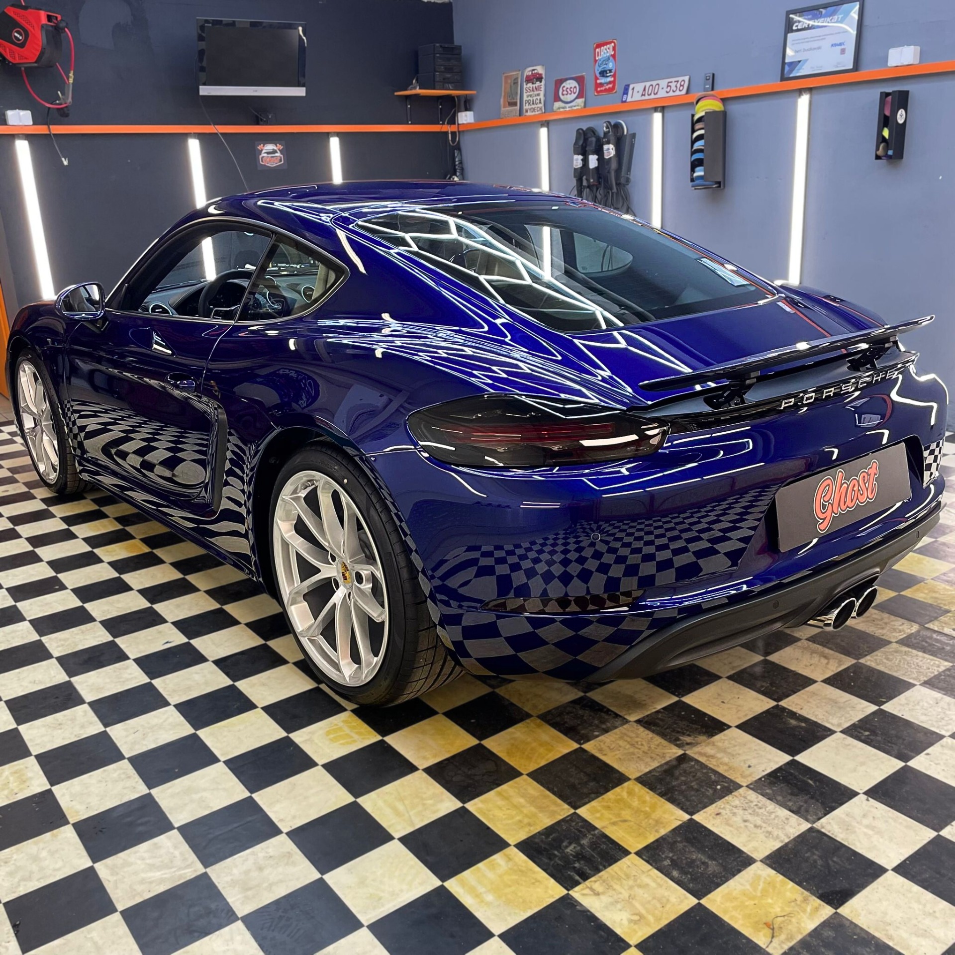 Błyszczący, granatowy Porsche 718 Cayman po detailingu, widoczne odbicia szachownicy posadzki w lakierze, logo firmy detailingowej na tylnym zderzaku.