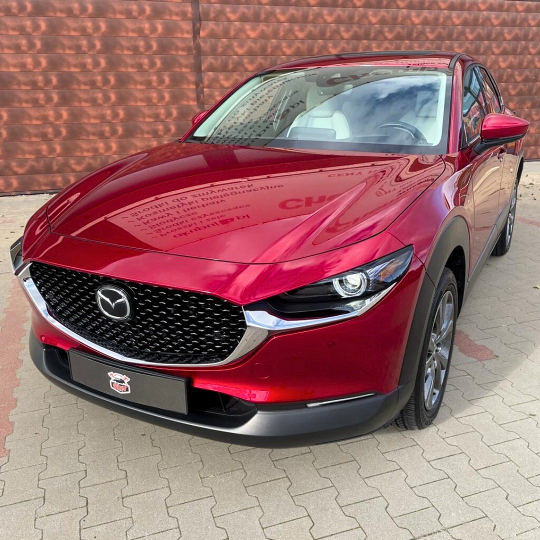 Błyszcząca, czerwona Mazda CX-30 po zabiegach detailingowych, odbijająca w lakierze otoczenie, zaparkowana na brukowanej nawierzchni przed brązową ścianą.