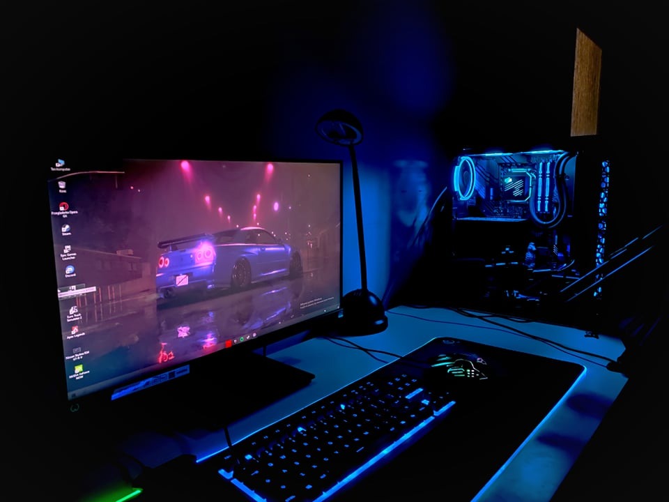 Komputer stacjonarny z podświetleniem RGB, monitor wyświetlający grafikę samochodu, klawiatura i mysz z podświetleniem, całość w ciemnym pomieszczeniu z niebieską poświatą.