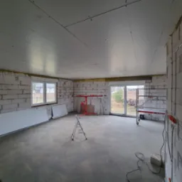 Wnętrze surowego domu w trakcie prac wykończeniowych: ściany z betonu komórkowego, zamontowane płyty gipsowo-kartonowe na suficie, widoczne przewody elektryczne, drabina i podnośnik do płyt.