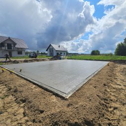 WWS CHOJNICE SPÓŁKA Z OGRANICZONĄ ODPOWIEDZIALNOŚCIĄ - Wylana, świeża płyta fundamentowa z widoczną izolacją obwodową styropianem, na piaszczystym gruncie, w tle dwa domy w różnym stanie budowy oraz błękitne niebo z chmurami.