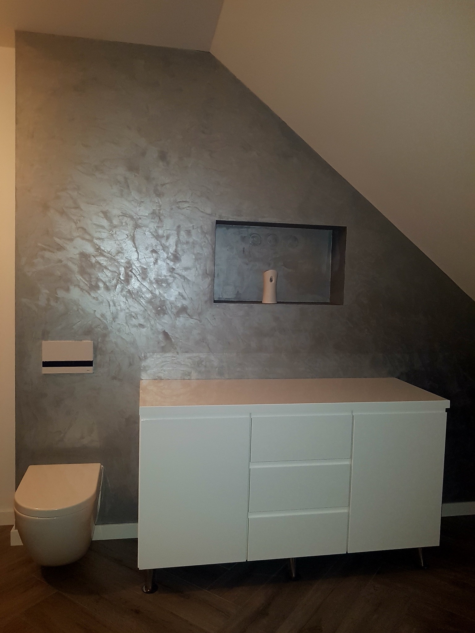 Nowoczesna łazienka na poddaszu z dekoracyjną, srebrzystą ścianą, zabudową meblową z białymi frontami i wiszącą toaletą.