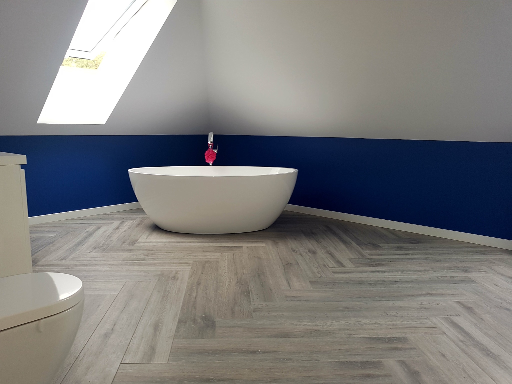 Nowoczesna łazienka na poddaszu z wolnostojącą białą wanną, drewnopodobną podłogą ułożoną we wzór jodełki i oknem dachowym.