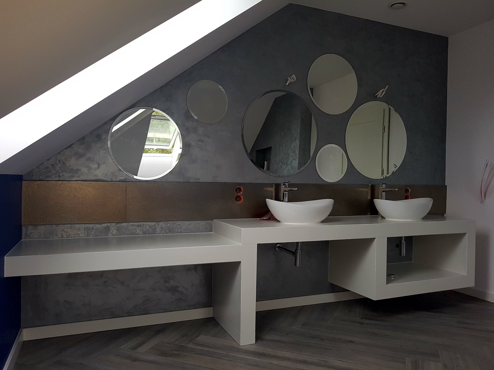 Nowoczesna łazienka na poddaszu z szarą ścianą, okrągłymi lustrami, białymi umywalkami nablatowymi i blatem z szafkami, podłoga w jodełkę.