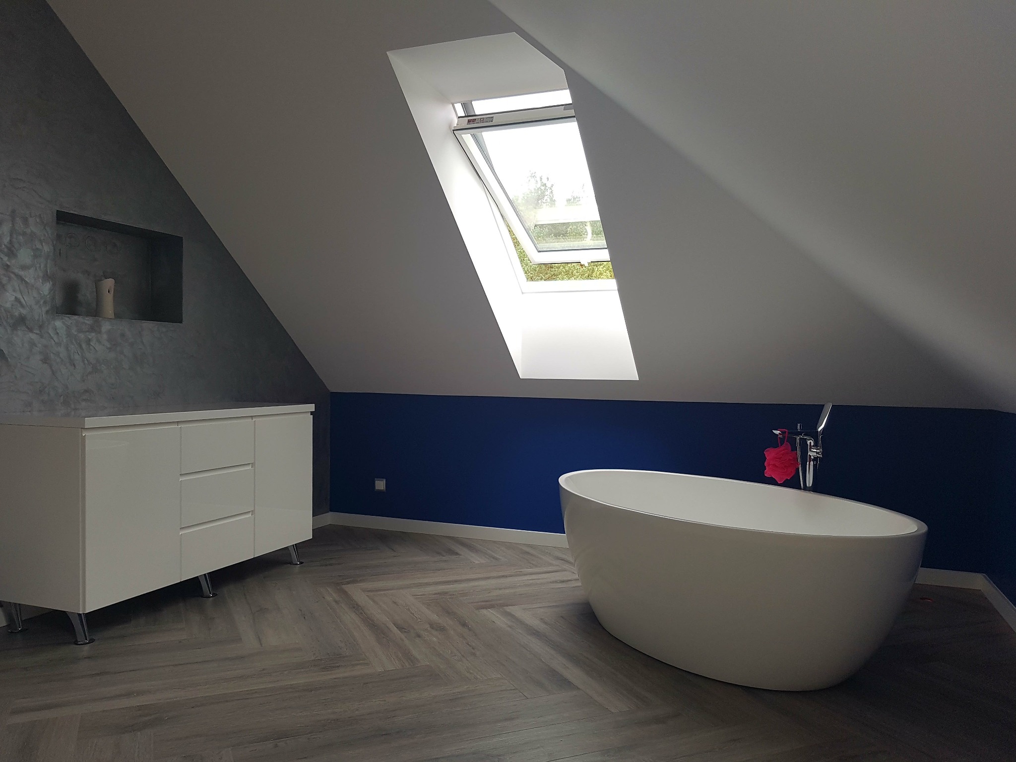 Nowoczesna łazienka na poddaszu z wolnostojącą wanną, białą komodą, szarą ścianą z wnęką i podłogą ułożoną w jodełkę, oświetlona naturalnym światłem z okna dachowego.