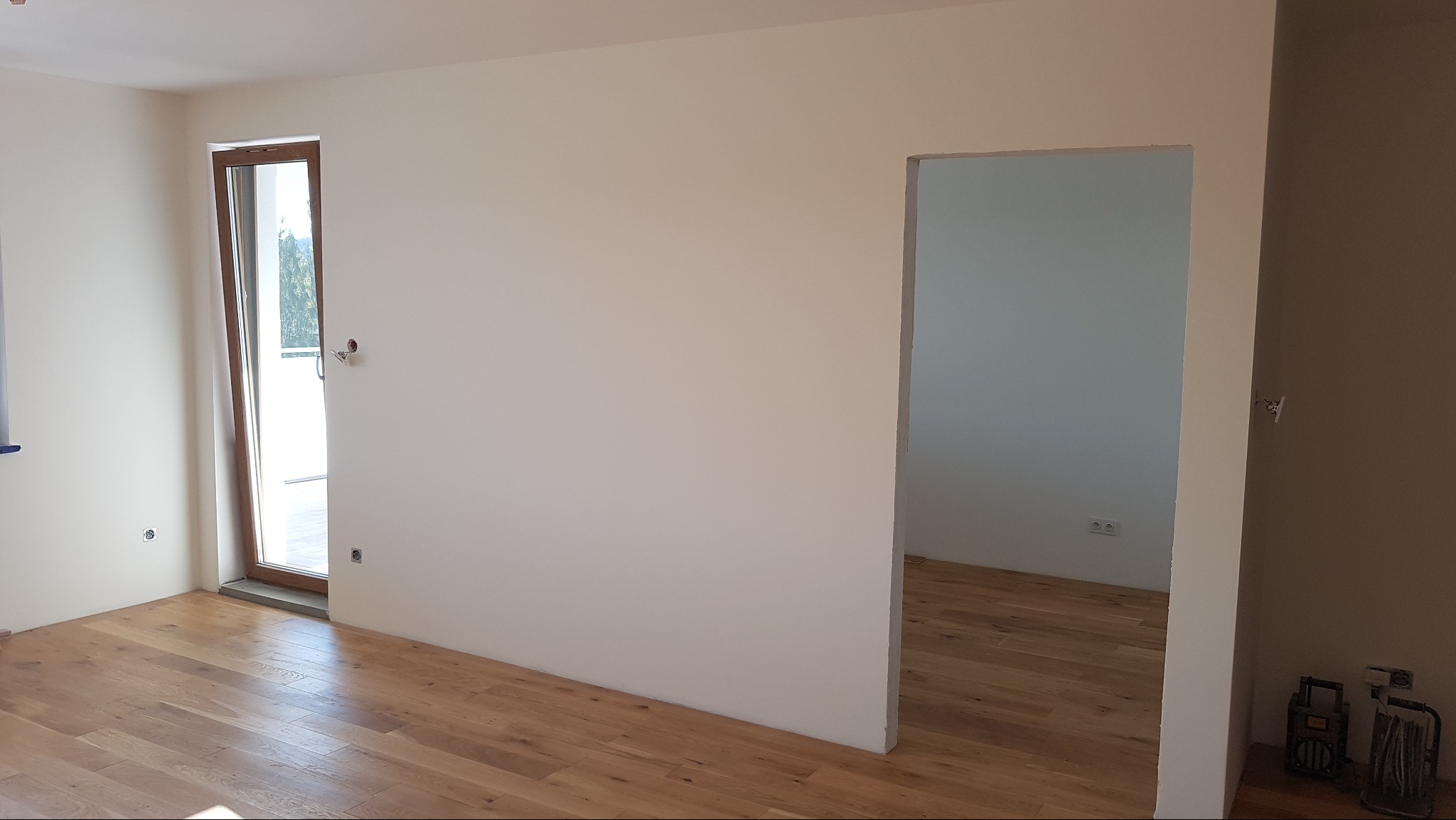 Wnętrze wykończonego pokoju z drewnianą podłogą, świeżo pomalowanymi ścianami na kremowo i otwartym przejściem do kolejnego pomieszczenia; widoczne gniazdka elektryczne i częściowo otwarte drzwi...