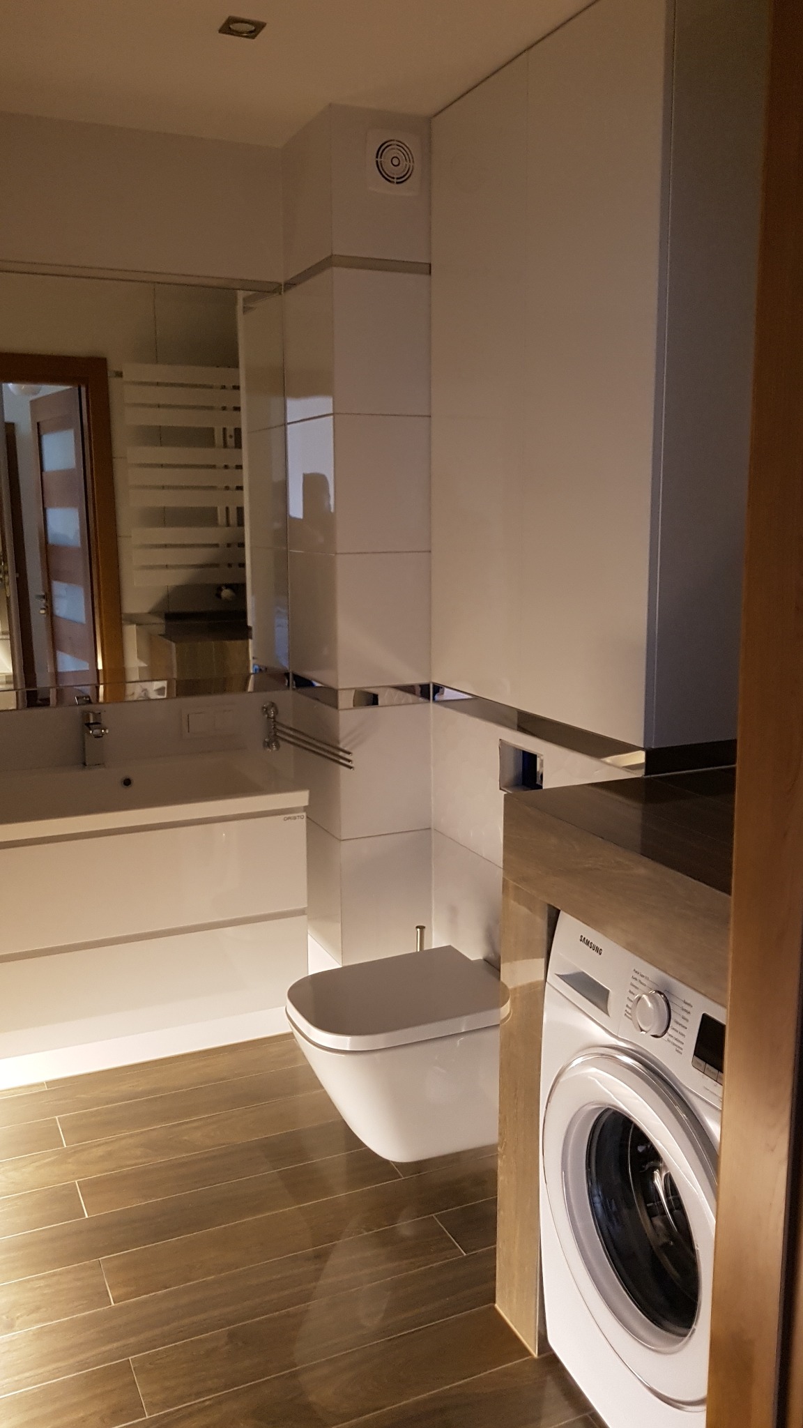 Nowoczesna łazienka z białymi meblami, toaletą podwieszaną i pralką Samsung, drewnopodobne płytki na podłodze, widoczny fragment lustra i wentylatora sufitowego.