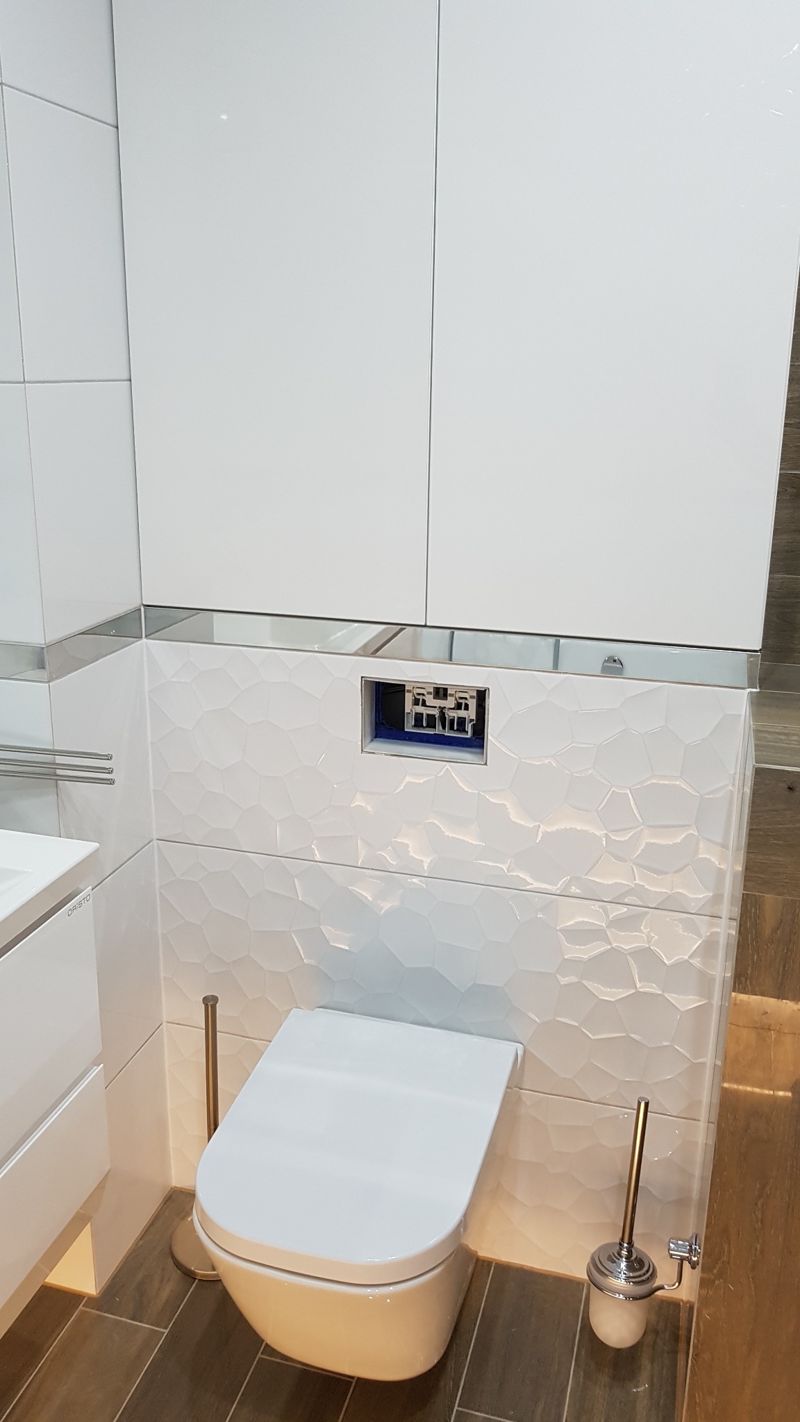 Nowoczesna toaleta podwieszana z białą miską i ukrytym stelażem, obudowana płytkami o geometrycznym wzorze, uzupełniona o białe szafki wiszące i akcesoria w kolorze szczotkowanego złota.