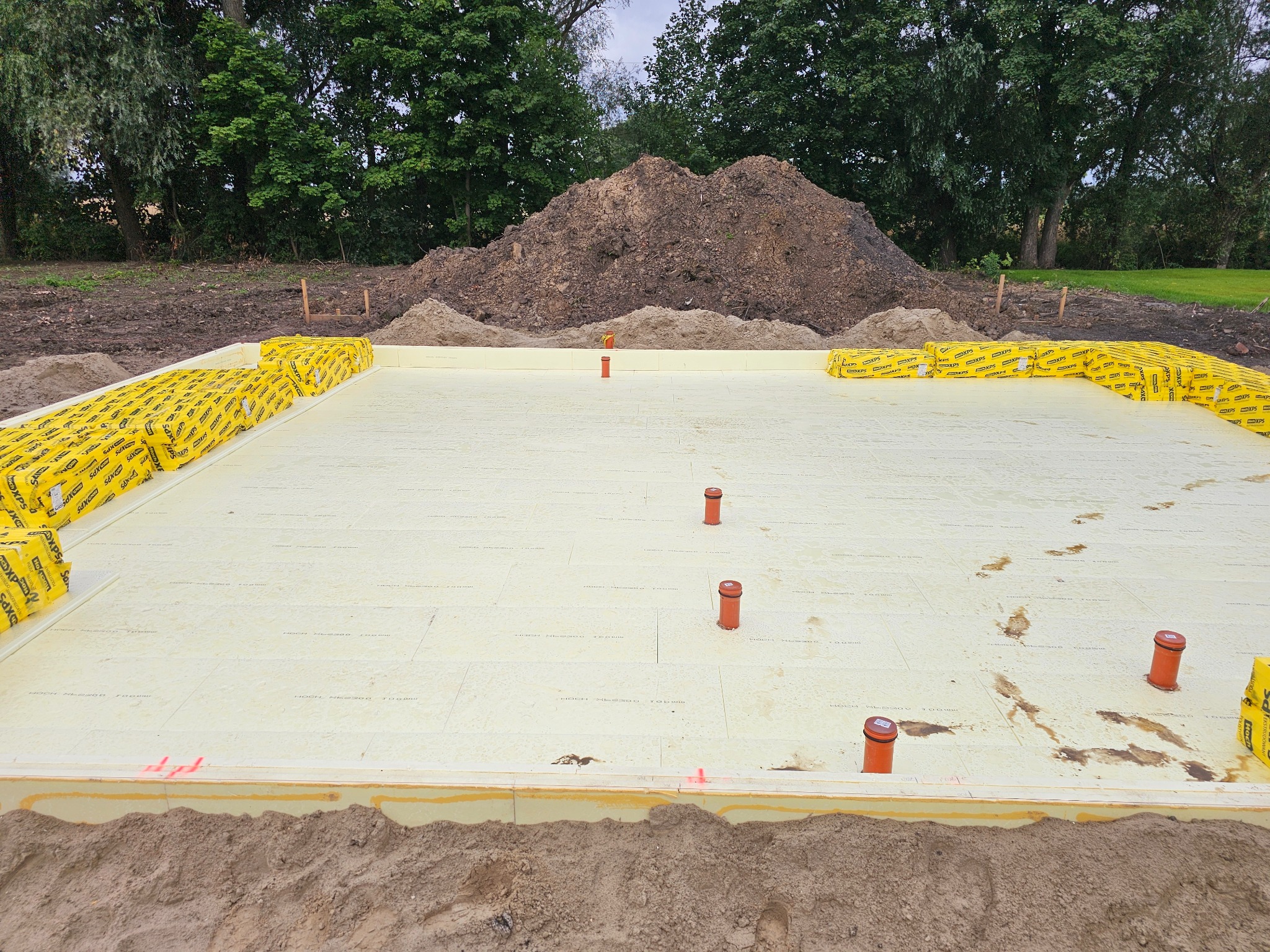 Płyta fundamentowa z izolacją termiczną XPS i pionowymi rurami kanalizacyjnymi, obłożona żółtymi paczkami izolacji, z hałdą ziemi w tle.