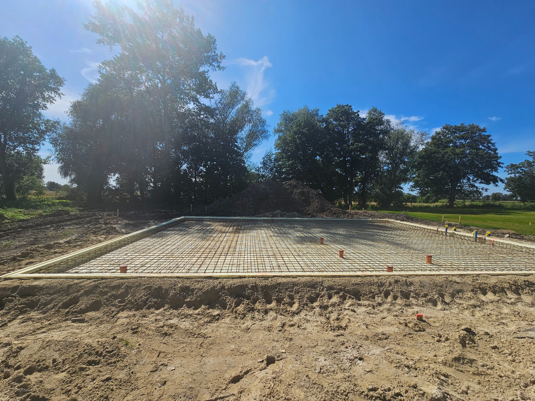 Przygotowany zbrojony szalunek pod płytę fundamentową na piaszczystym gruncie, widoczne wystające rury instalacyjne, w tle drzewa i błękitne niebo.