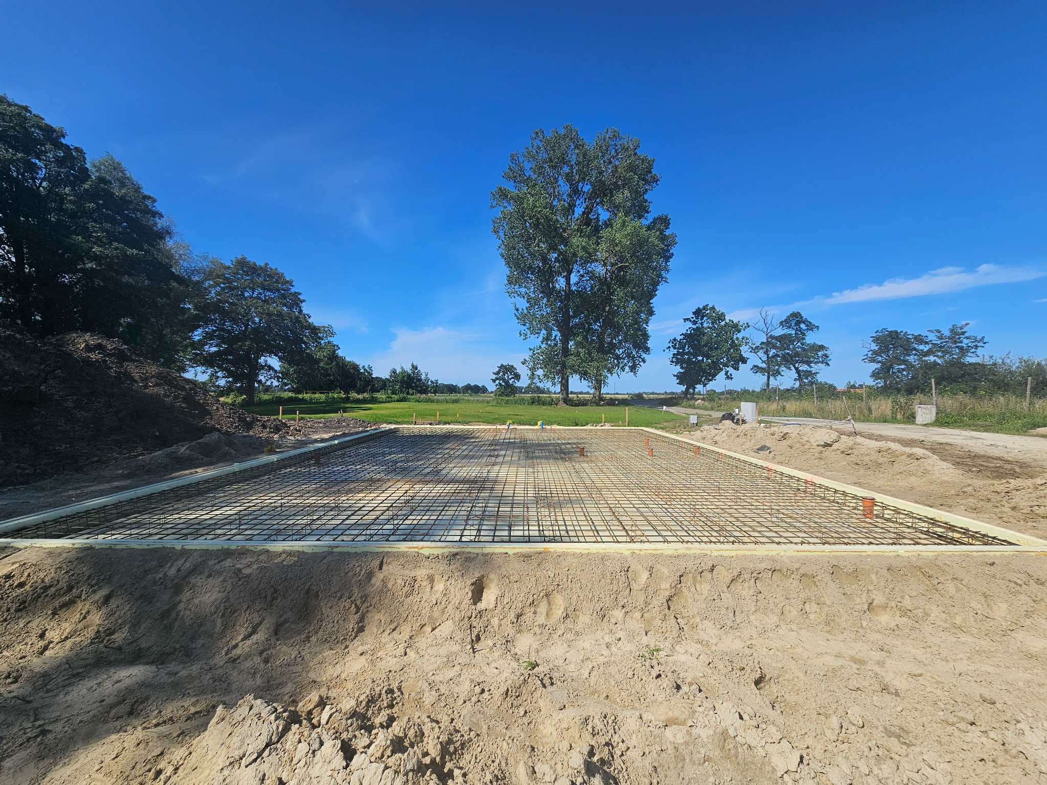 Przygotowany zbrojony fundament pod budowę domu, widoczne szalunki z izolacją termiczną, na tle błękitnego nieba i zieleni drzew.