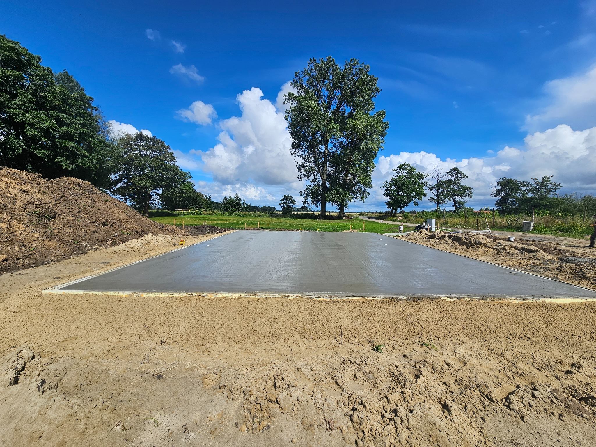 Świeżo wylana, gładka płyta fundamentowa otoczona piaskiem i z widokiem na drzewa i błękitne niebo z chmurami.