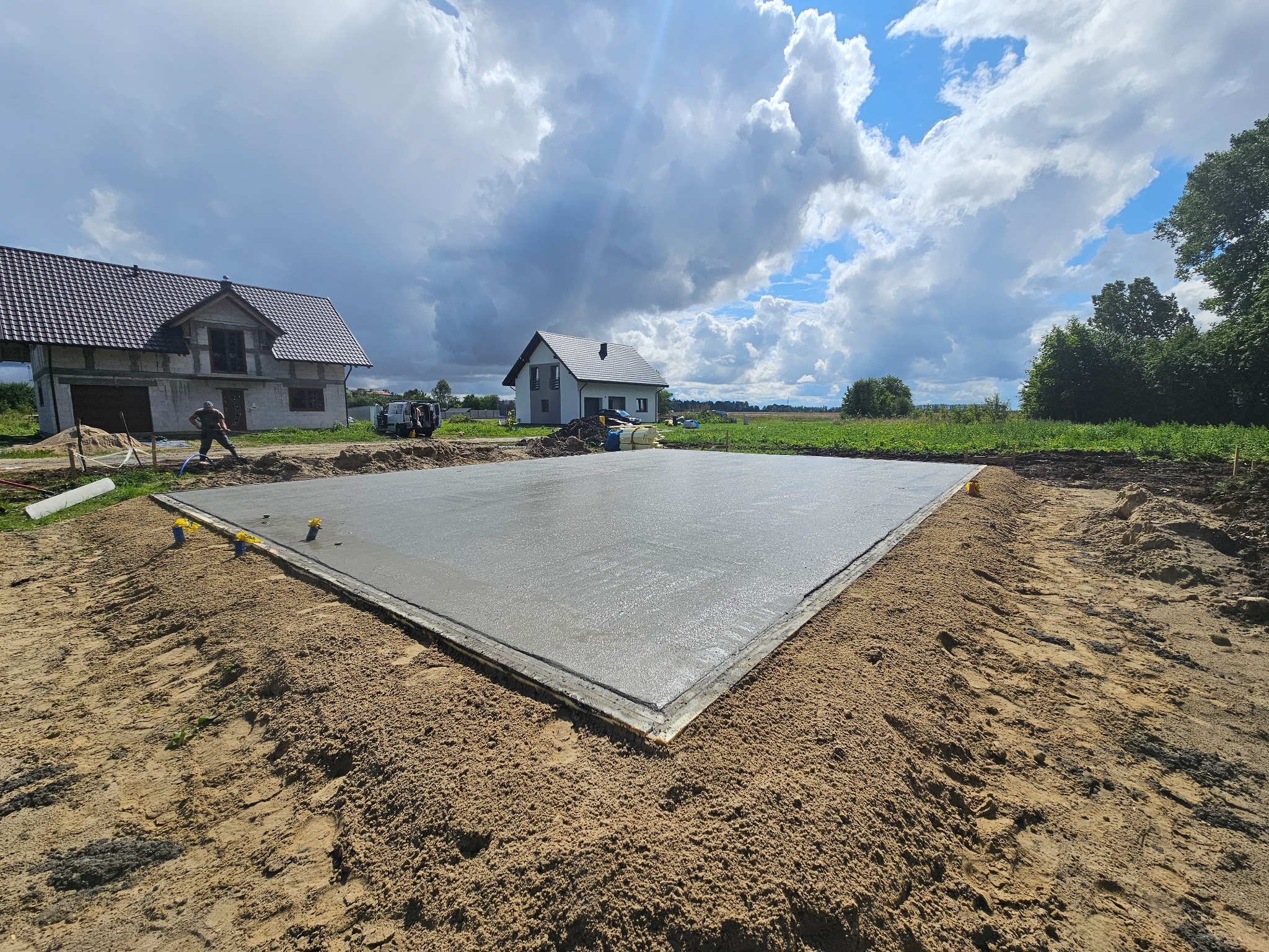Wylana, świeża płyta fundamentowa z widoczną izolacją obwodową styropianem, na piaszczystym gruncie, w tle dwa domy w różnym stanie budowy oraz błękitne niebo z chmurami.