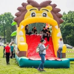Dmuchana zjeżdżalnia w kształcie lwa z dziećmi na trawniku, otoczona taśmą ostrzegawczą i osobami w tle, w słoneczny dzień.