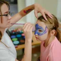 Artystka maluje twarz dziecka farbami, tworząc wzór w odcieniach niebieskiego i różowego. Widoczny paleta kolorów i pędzel. Dziewczynka ma na sobie różową koszulkę.