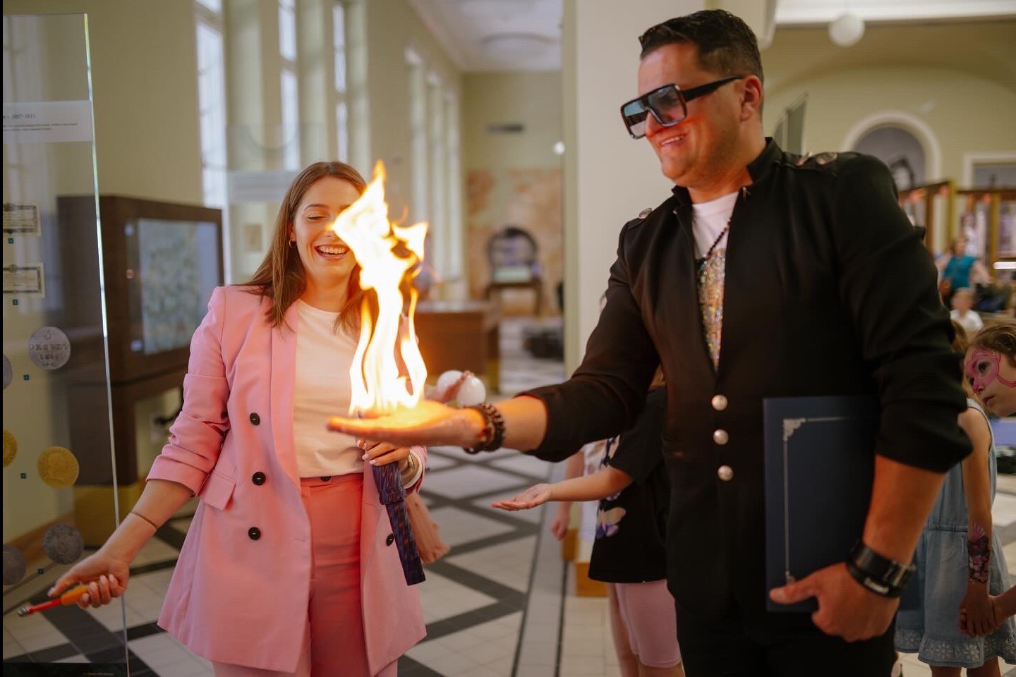 Animator w czarnym fraku prezentuje płomień na dłoni, rozbawiona kobieta w różowym garniturze obserwuje pokaz w eleganckim wnętrzu.