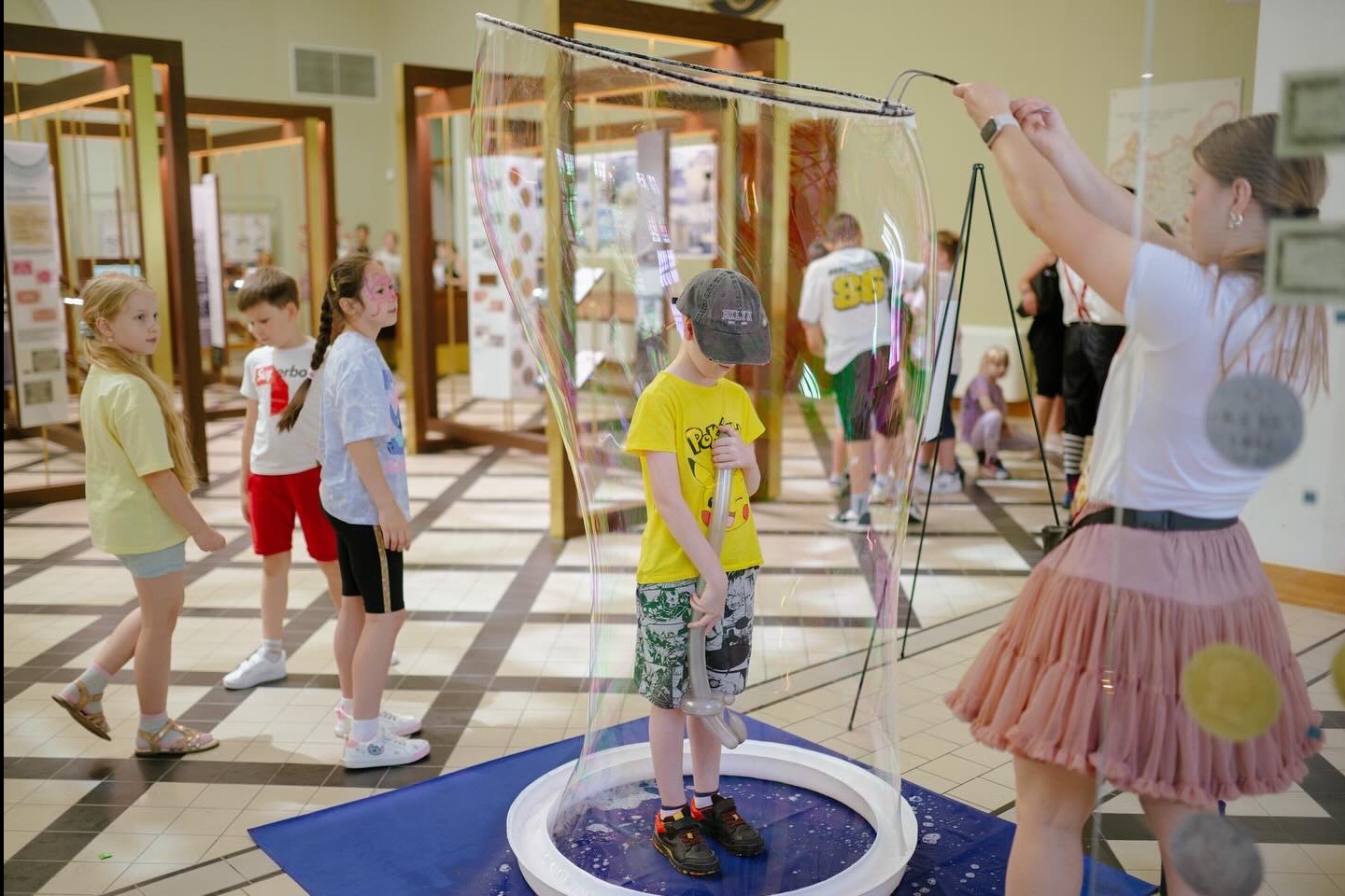 Chłopiec w gigantycznej bańce mydlanej, obsługa w tle, dzieci obserwują. Atrakcja eventowa w Krakowie, zabawa w pomieszczeniu, światło dzienne.