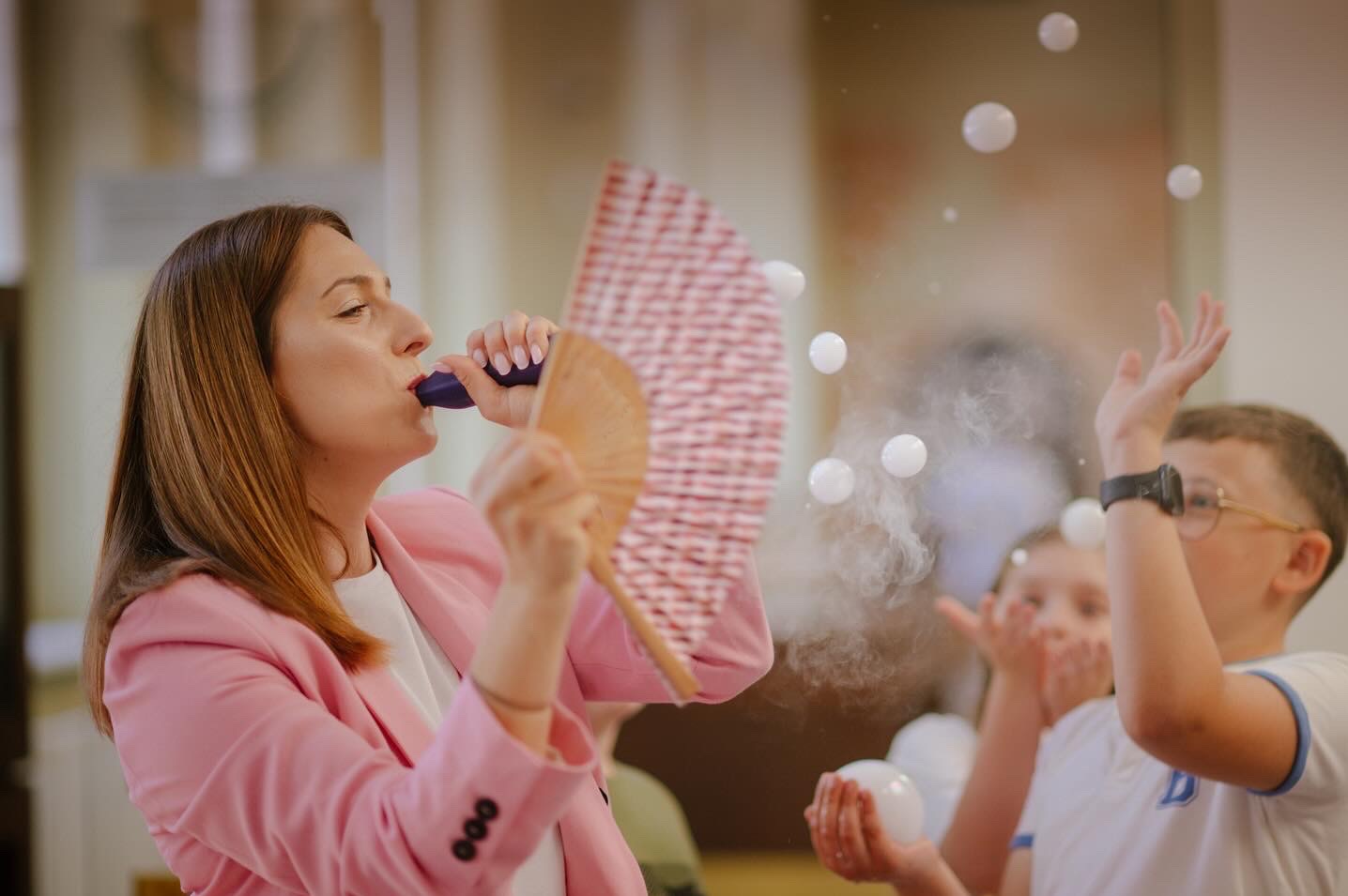 Animator dmucha bańki mydlane, trzymając wachlarz, dzieci próbują je łapać. Kobieta w różowej marynarce podczas pokazu baniek mydlanych na imprezie.