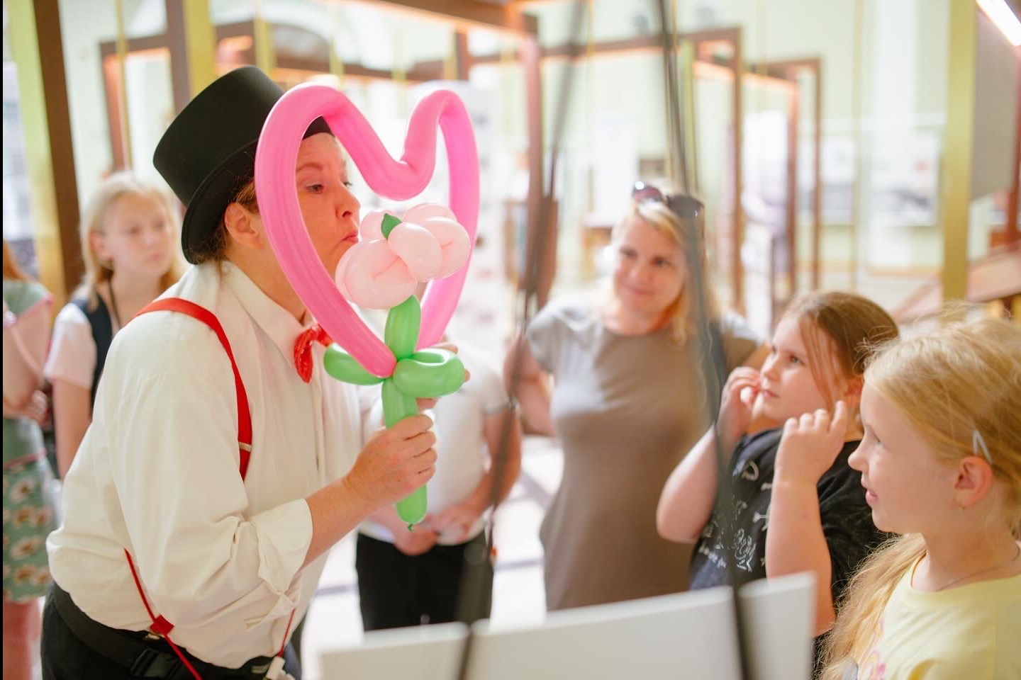 Animator w cylindrze tworzy balonową kompozycję kwiatu w sercu dla dzieci, w tle publiczność w jasnym pomieszczeniu.