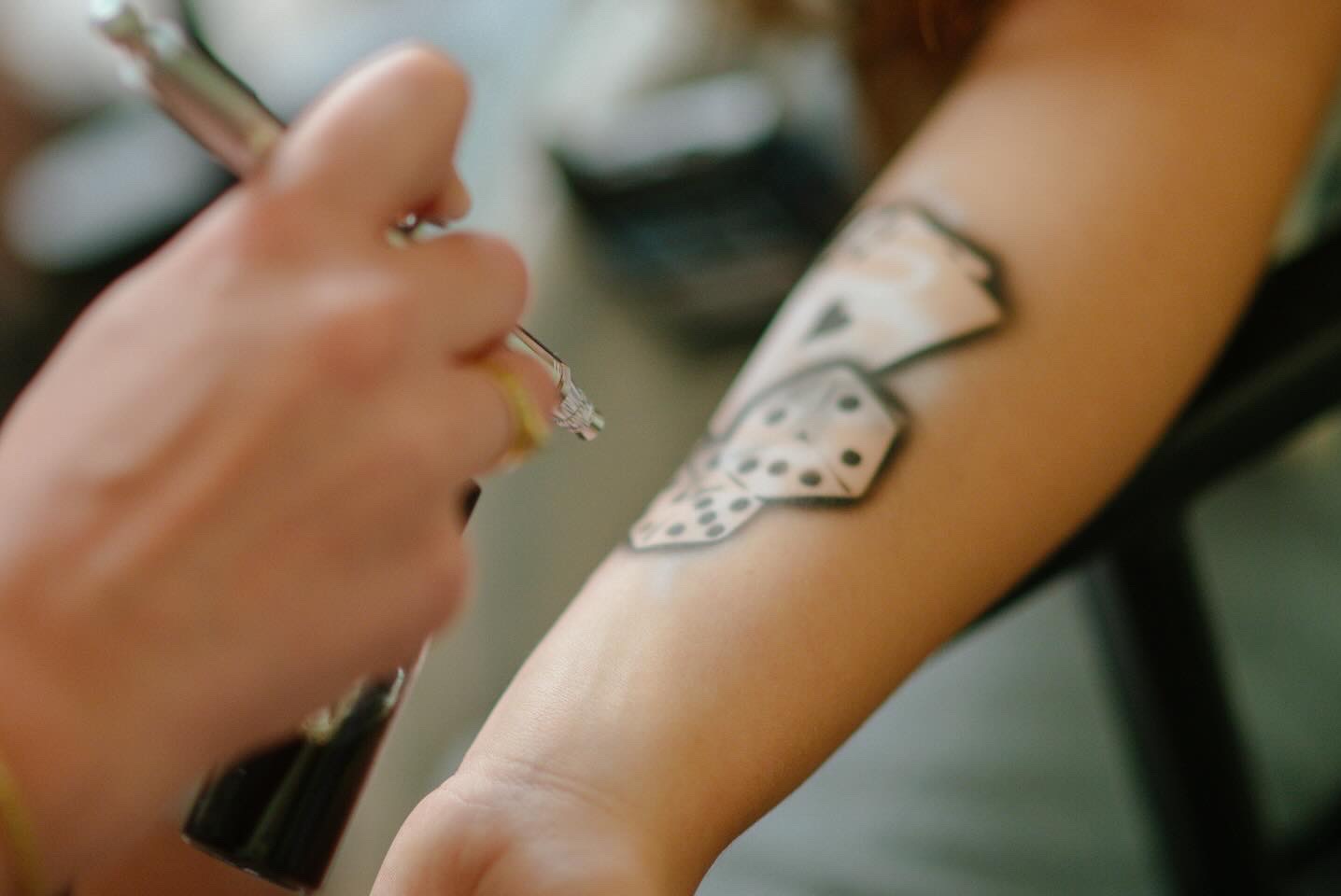 Aplikacja tymczasowego tatuażu aerografem na ramieniu, wzór kart i kostek do gry. Delikatne cieniowanie, bliska perspektywa, etap wykonywania.