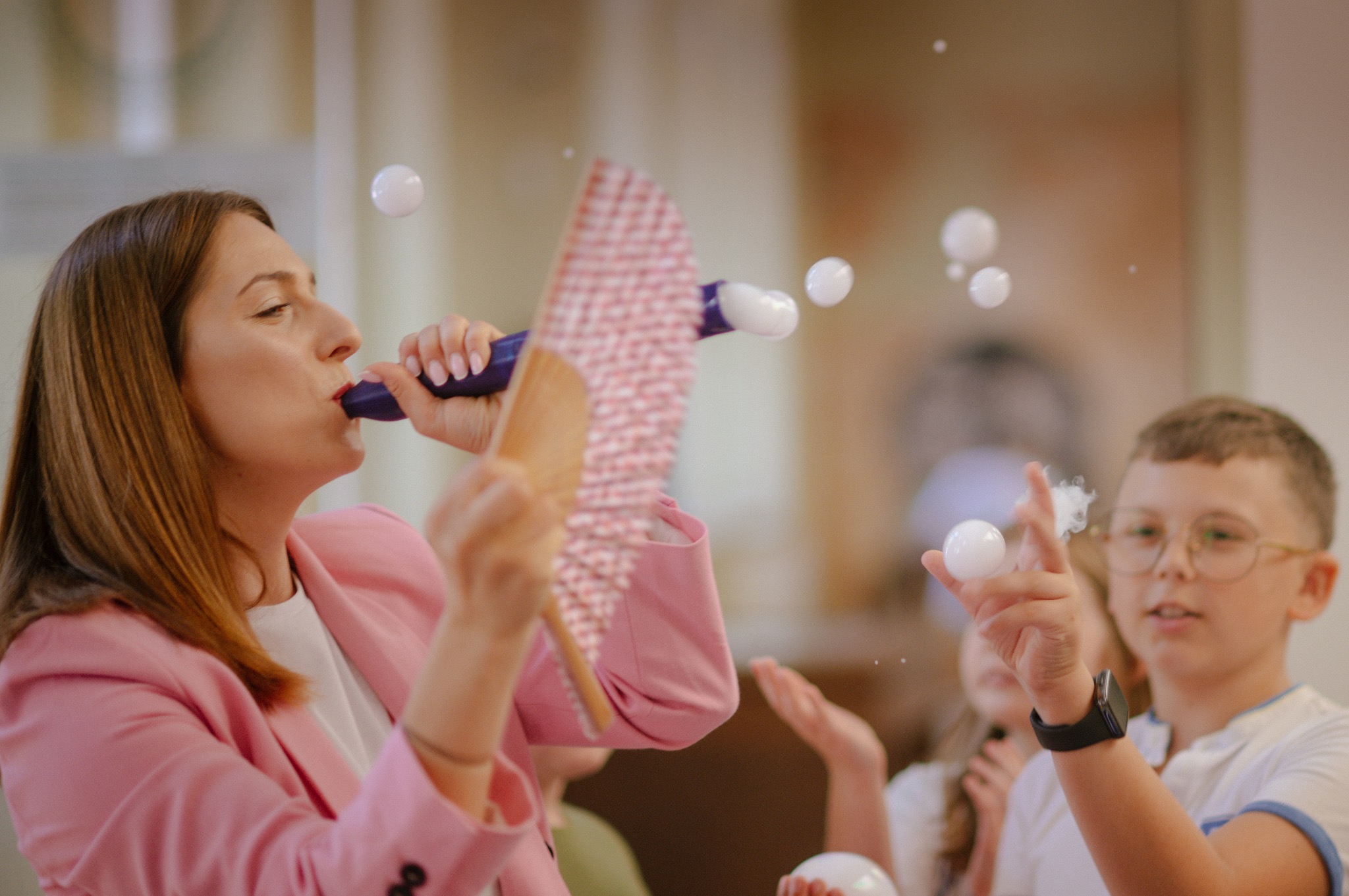 Animator w różowym żakiecie dmucha bańki mydlane, dzieci wyciągają ręce, by je złapać. Atrakcja na imprezie dla dzieci.