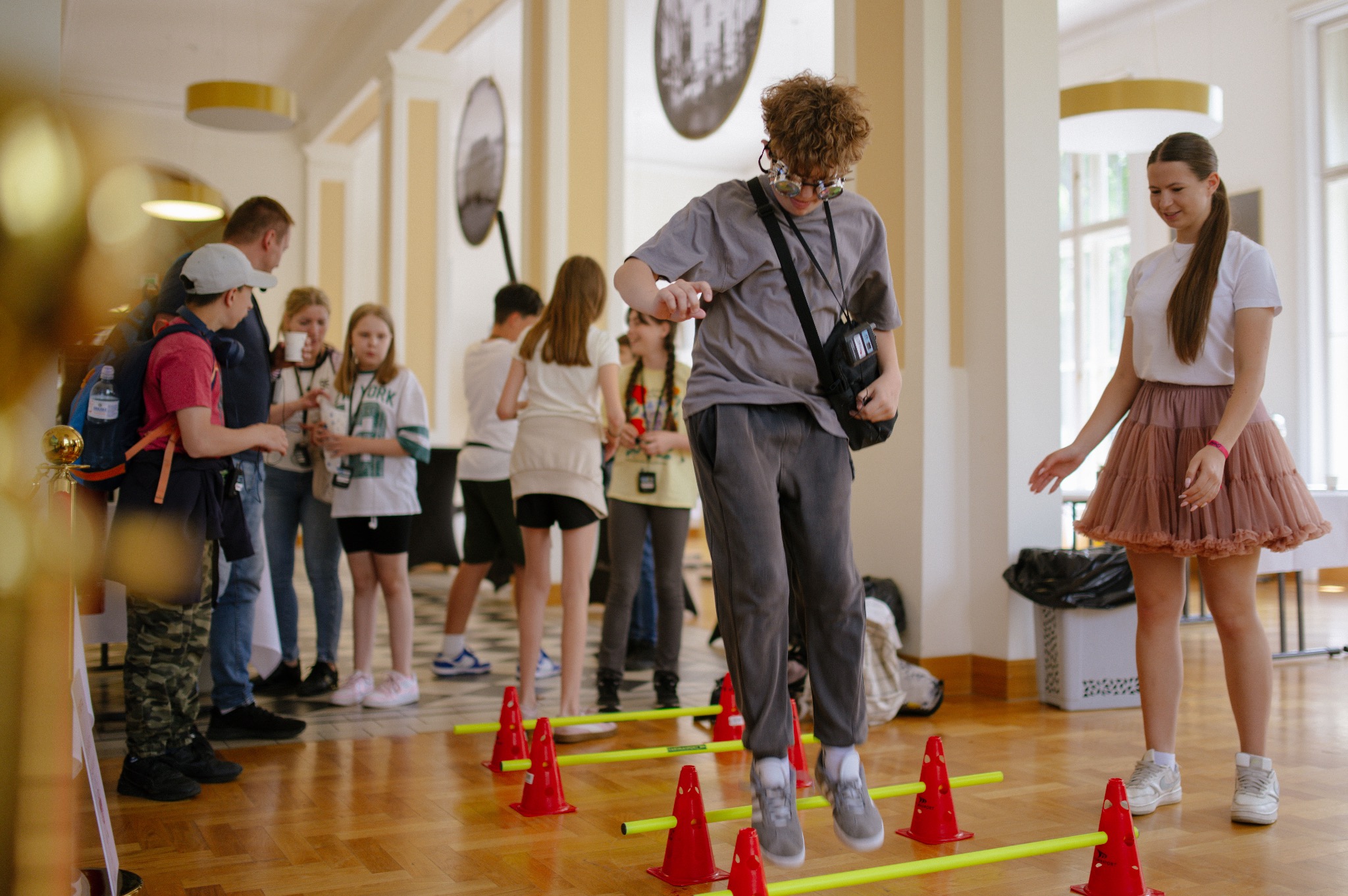 Chłopak w okularach skacze przez przeszkody na tle grupy dzieci i dorosłych w jasnym wnętrzu, prawdopodobnie podczas animacji lub eventu.