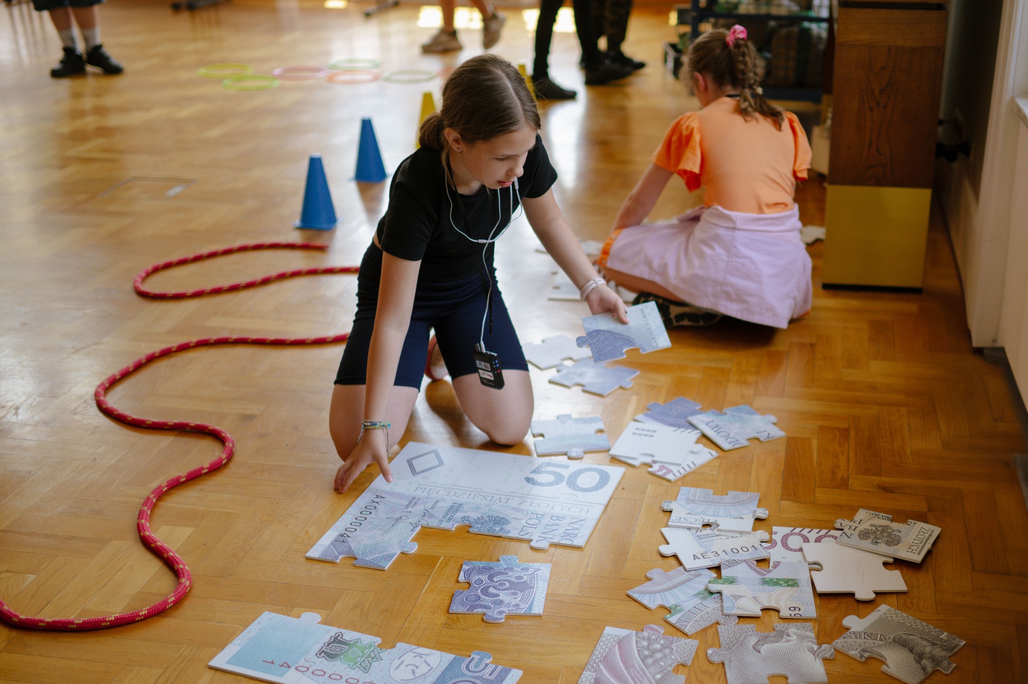 Dziewczynka układa puzzle z motywem banknotów na drewnianej podłodze, w tle widoczne inne dzieci i elementy wyposażenia sali zabaw.
