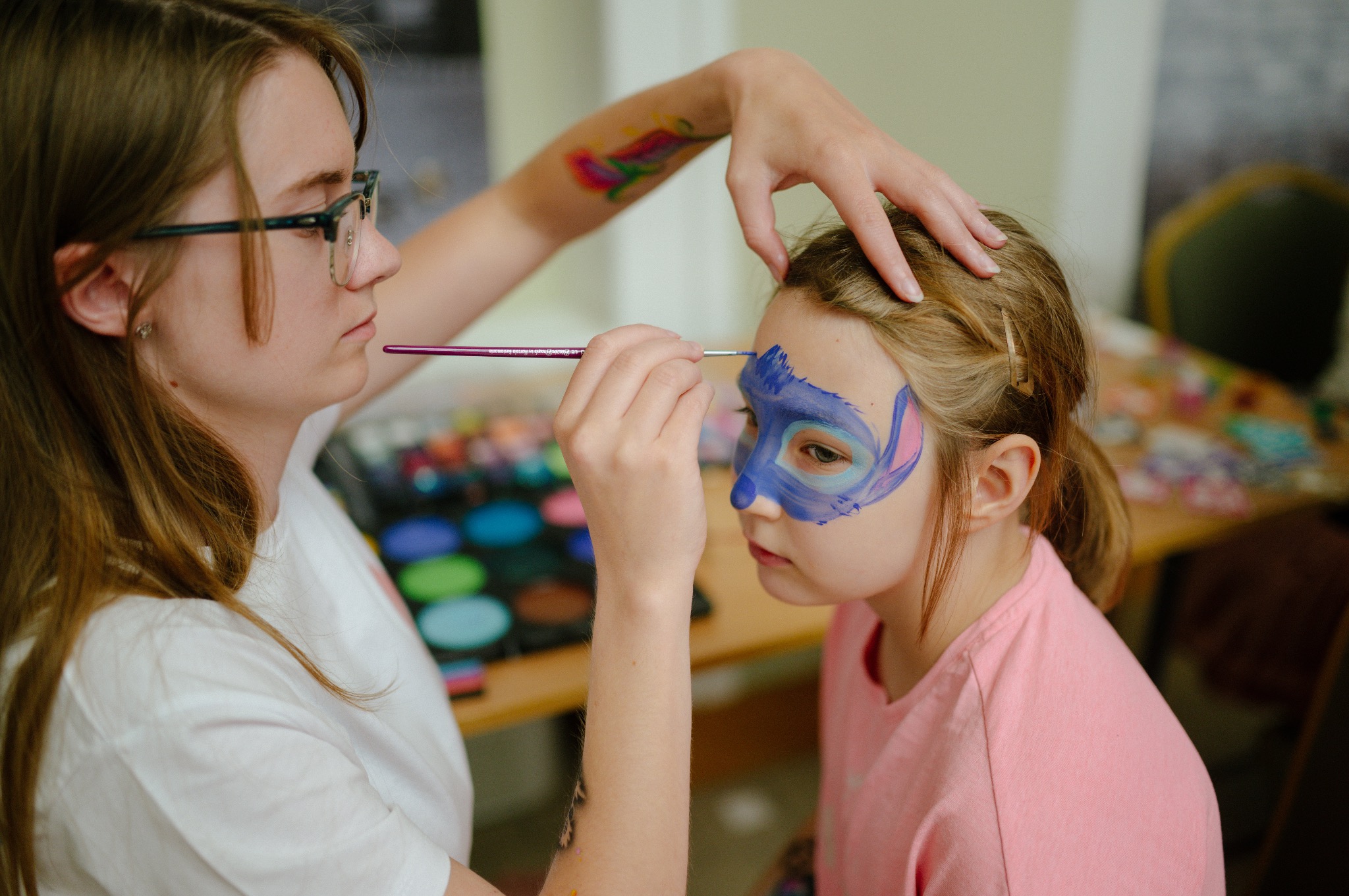Artystka maluje twarz dziecka farbami, tworząc wzór w odcieniach niebieskiego i różowego. Widoczny paleta kolorów i pędzel. Dziewczynka ma na sobie różową koszulkę.