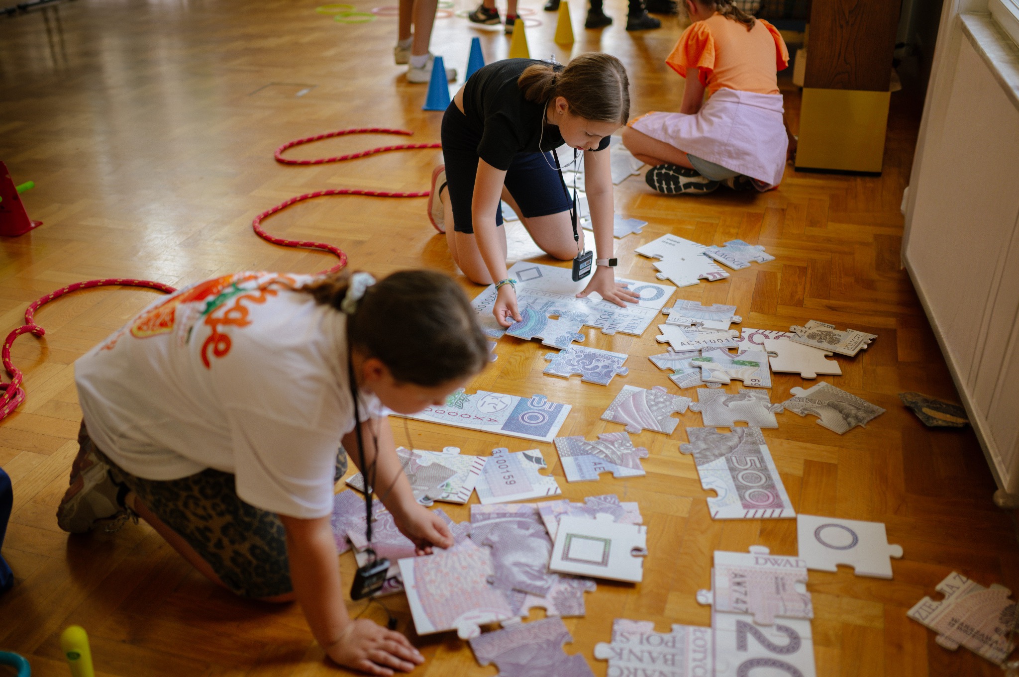 Dzieci układają na podłodze puzzle z motywem banknotów, otoczone kolorowymi rekwizytami, podczas animacji.