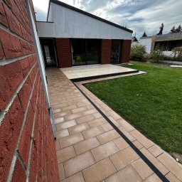Miłosz Kramarczyk Usługi Brukarskie - Ścieżka z beżowej kostki brukowej z czarnym obramowaniem prowadząca do nowoczesnego domu z czerwonej cegły i dużymi oknami, widok na zielony trawnik i częściowo ukończony budynek w tle.