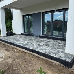 Miłosz Kramarczyk Usługi Brukarskie - Nowoczesny taras z szarej kostki brukowej obramowany czarnym obrzeżem, przylegający do budynku z dużymi oknami. Widoczne elementy wykończeniowe.