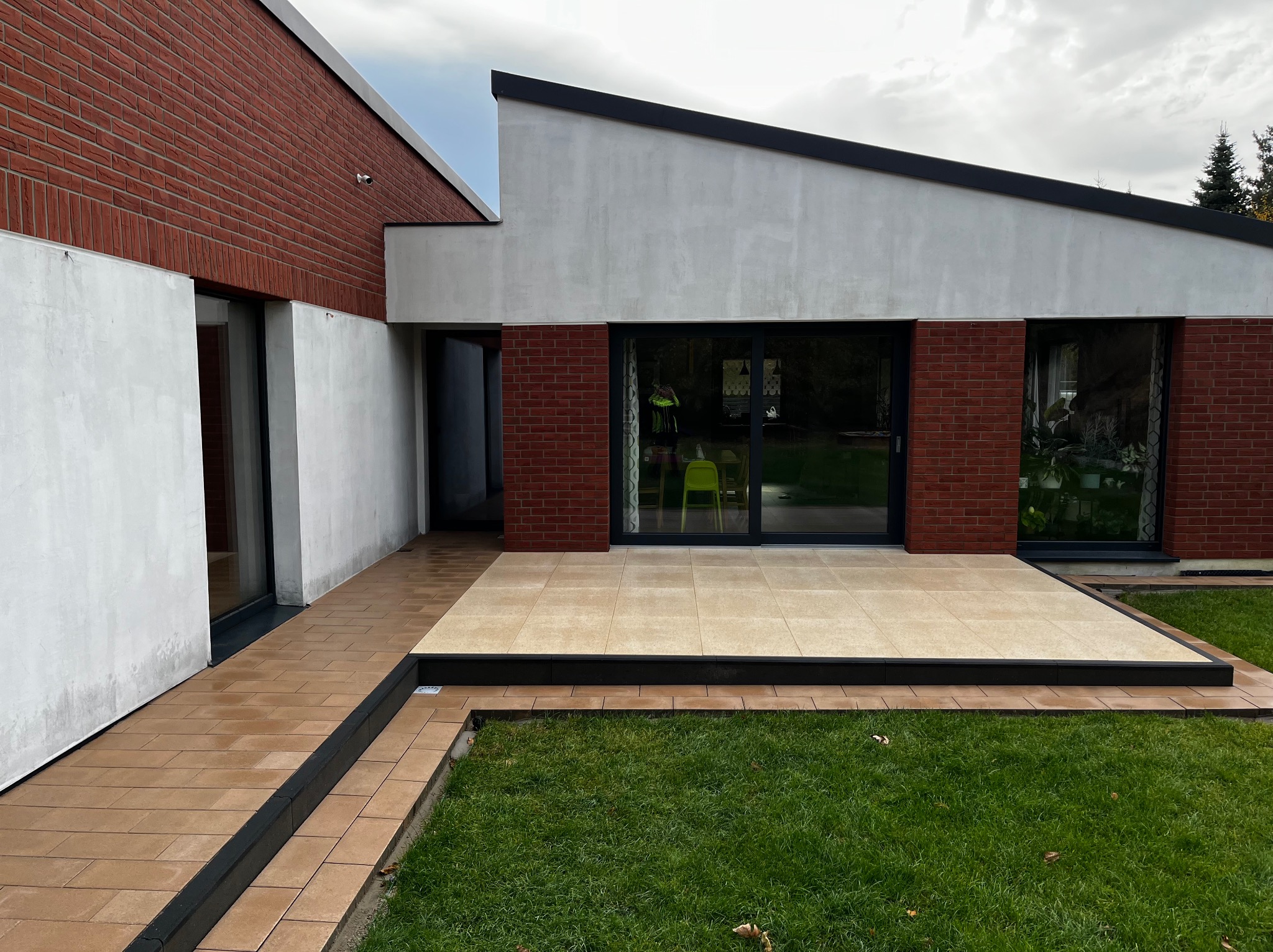 Nowoczesny taras z jasnych płyt, obrzeża z ciemnego kamienia, trawnik i elewacja budynku z cegły i białego tynku.