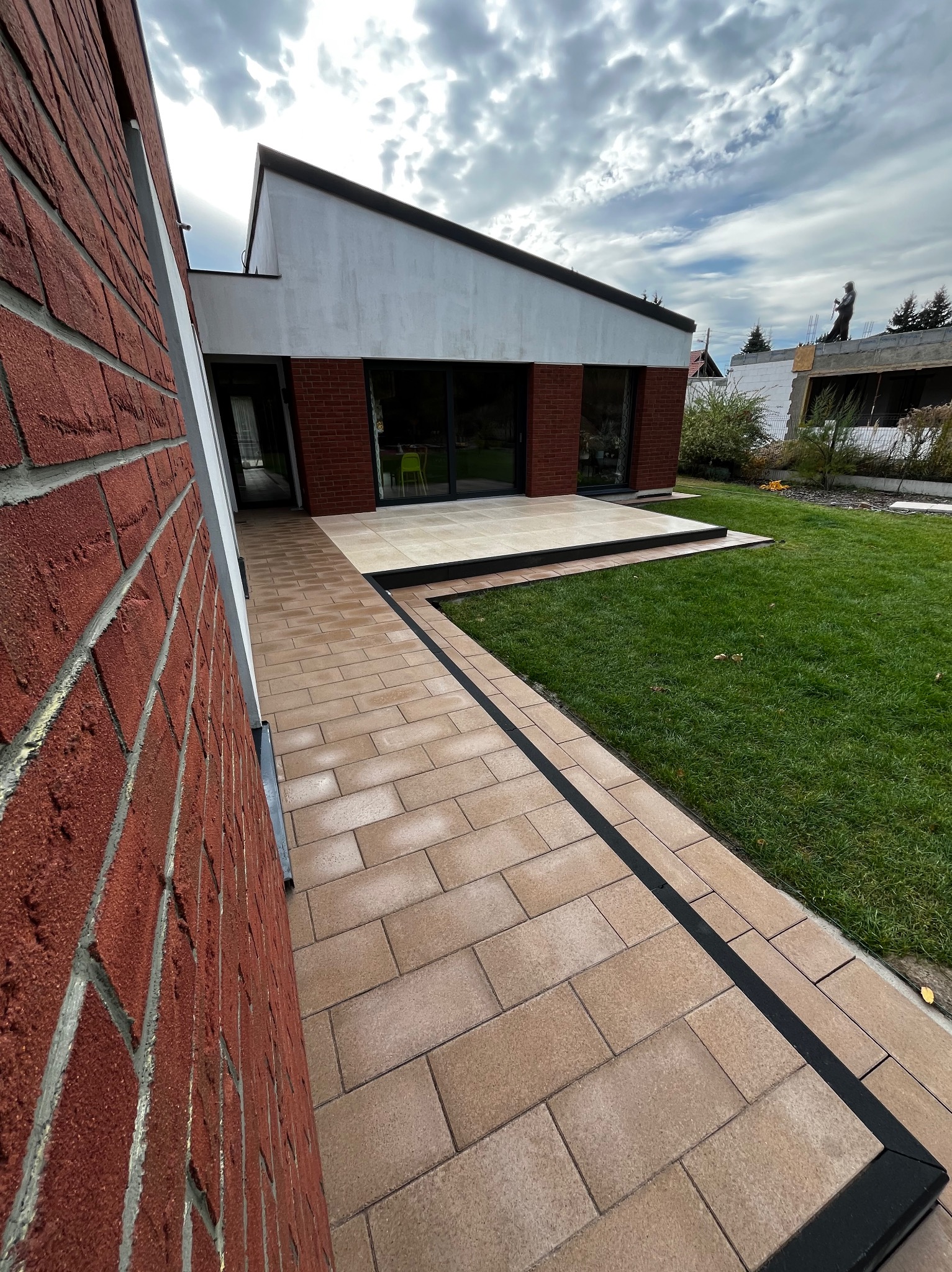 Ścieżka z beżowej kostki brukowej z czarnym obramowaniem prowadząca do nowoczesnego domu z czerwonej cegły i dużymi oknami, widok na zielony trawnik i częściowo ukończony budynek w tle.