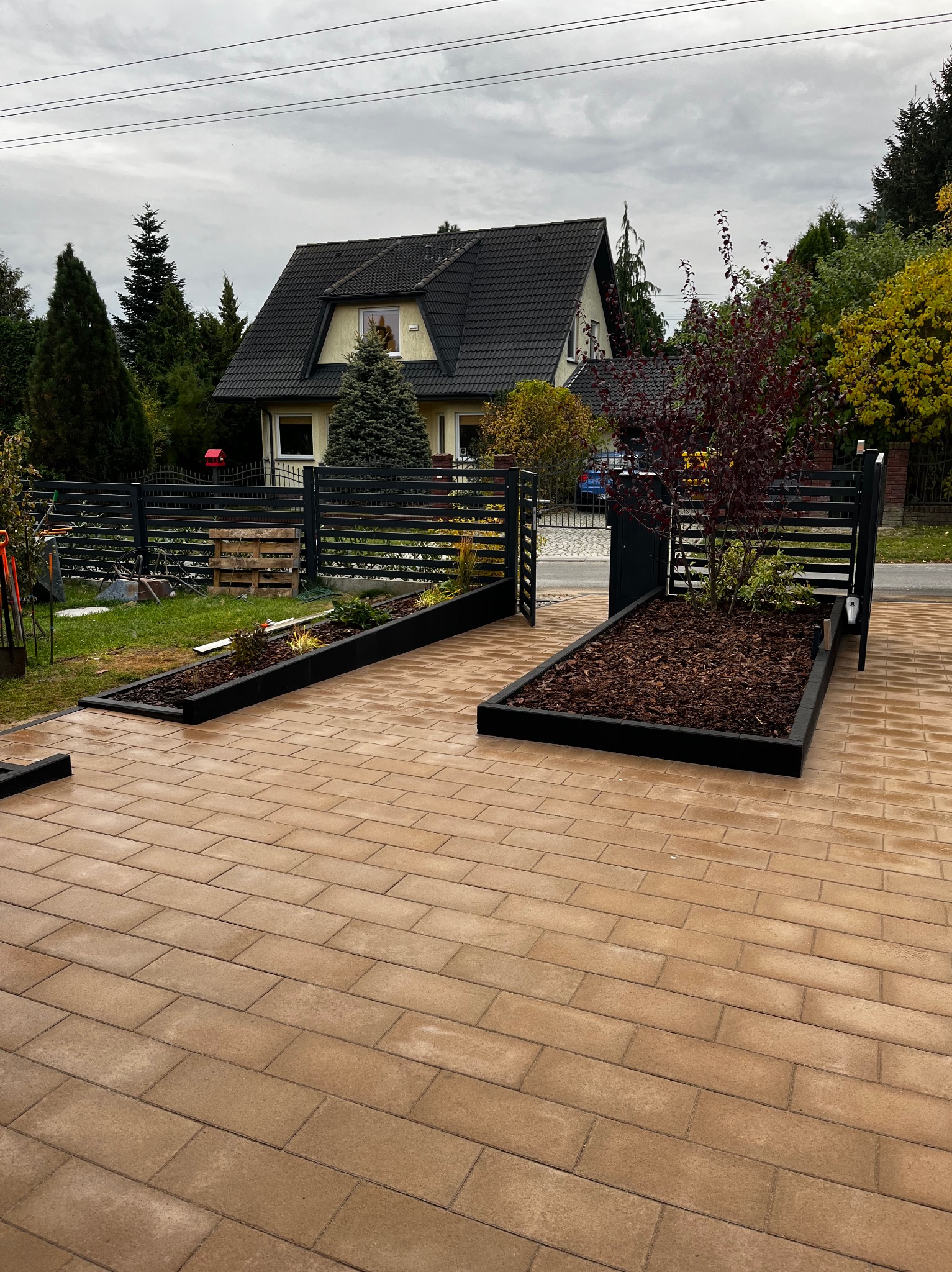 Ułożona kostka brukowa w odcieniach beżu prowadząca do nowoczesnego, czarnego ogrodzenia z dwoma podwyższonymi rabatami; jedna z korą, druga z roślinami.