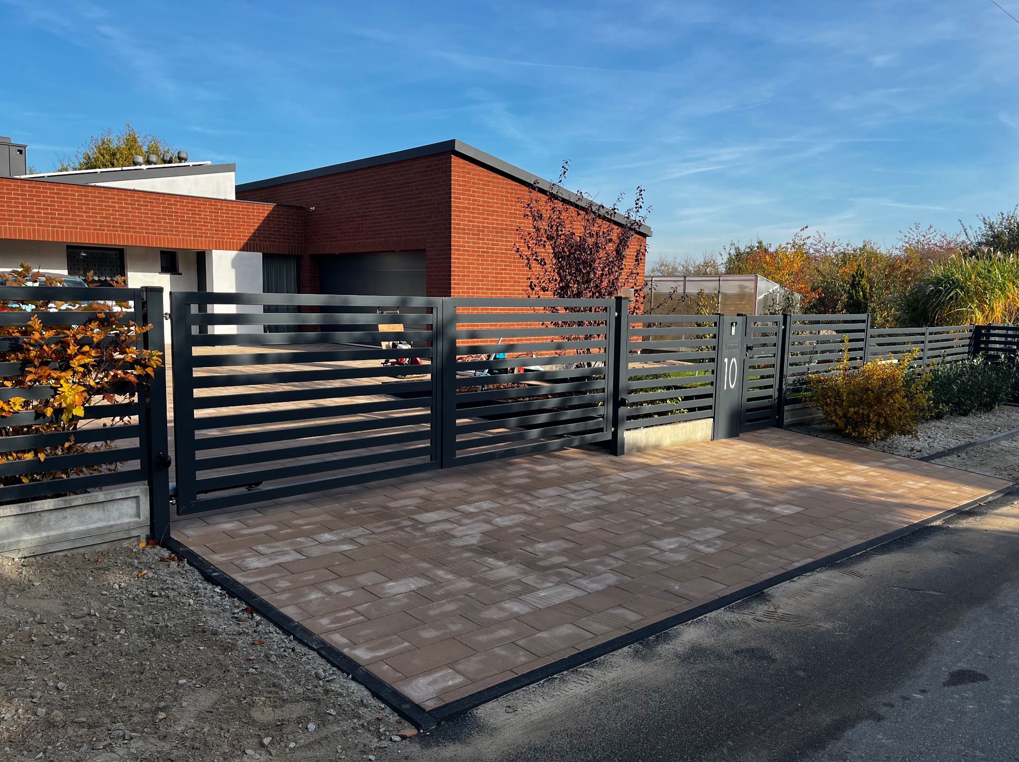Nowoczesne, poziome ogrodzenie panelowe w kolorze grafitowym z numerem domu '10', prowadzące do podjazdu wyłożonego brązową kostką brukową, na tle domu z czerwonej cegły i błękitnego nieba.