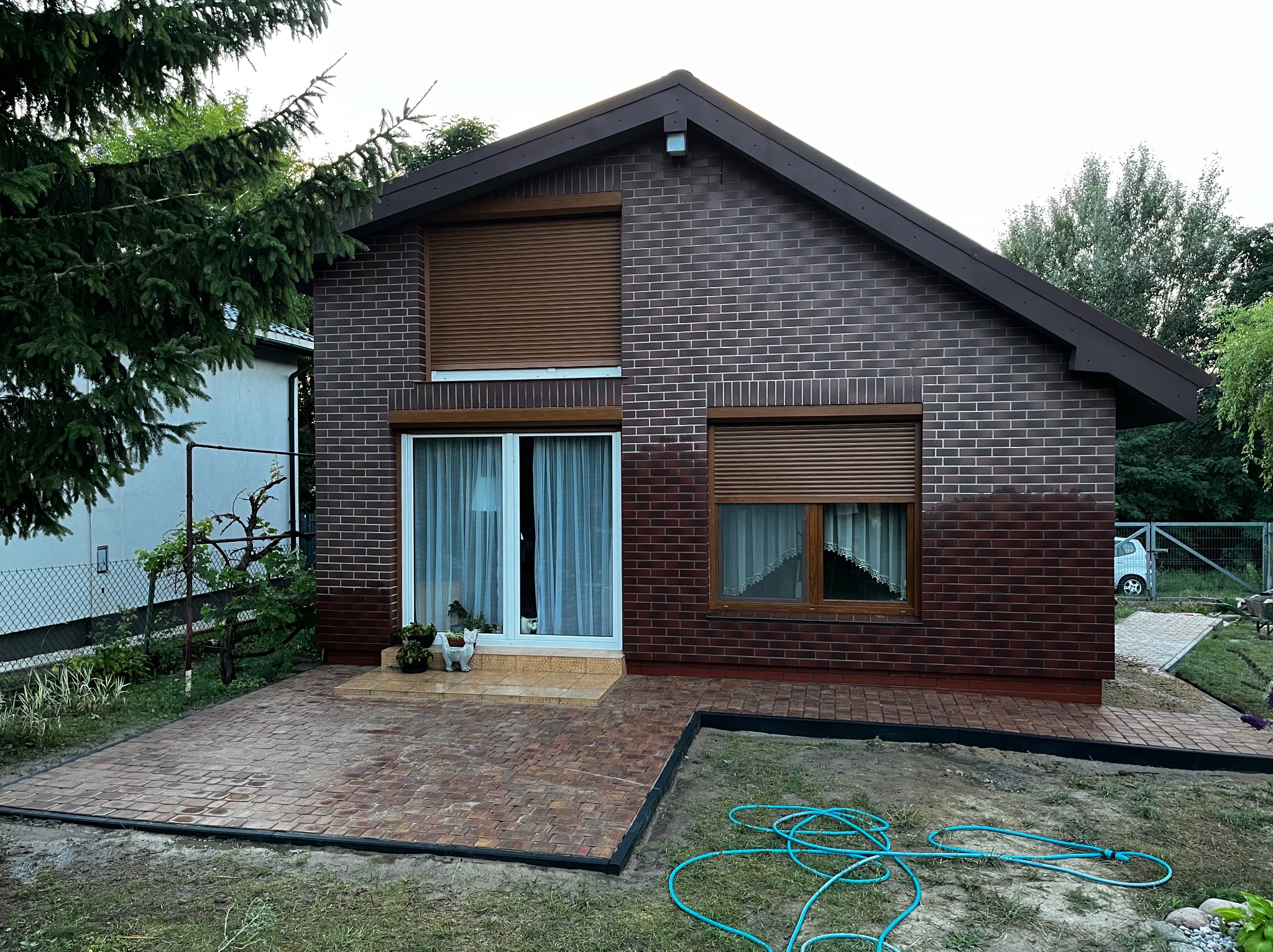 Nowo ułożona, brązowa kostka brukowa przed domem z cegły, widoczny fragment trawnika z wężem ogrodowym, okna z roletami zewnętrznymi.