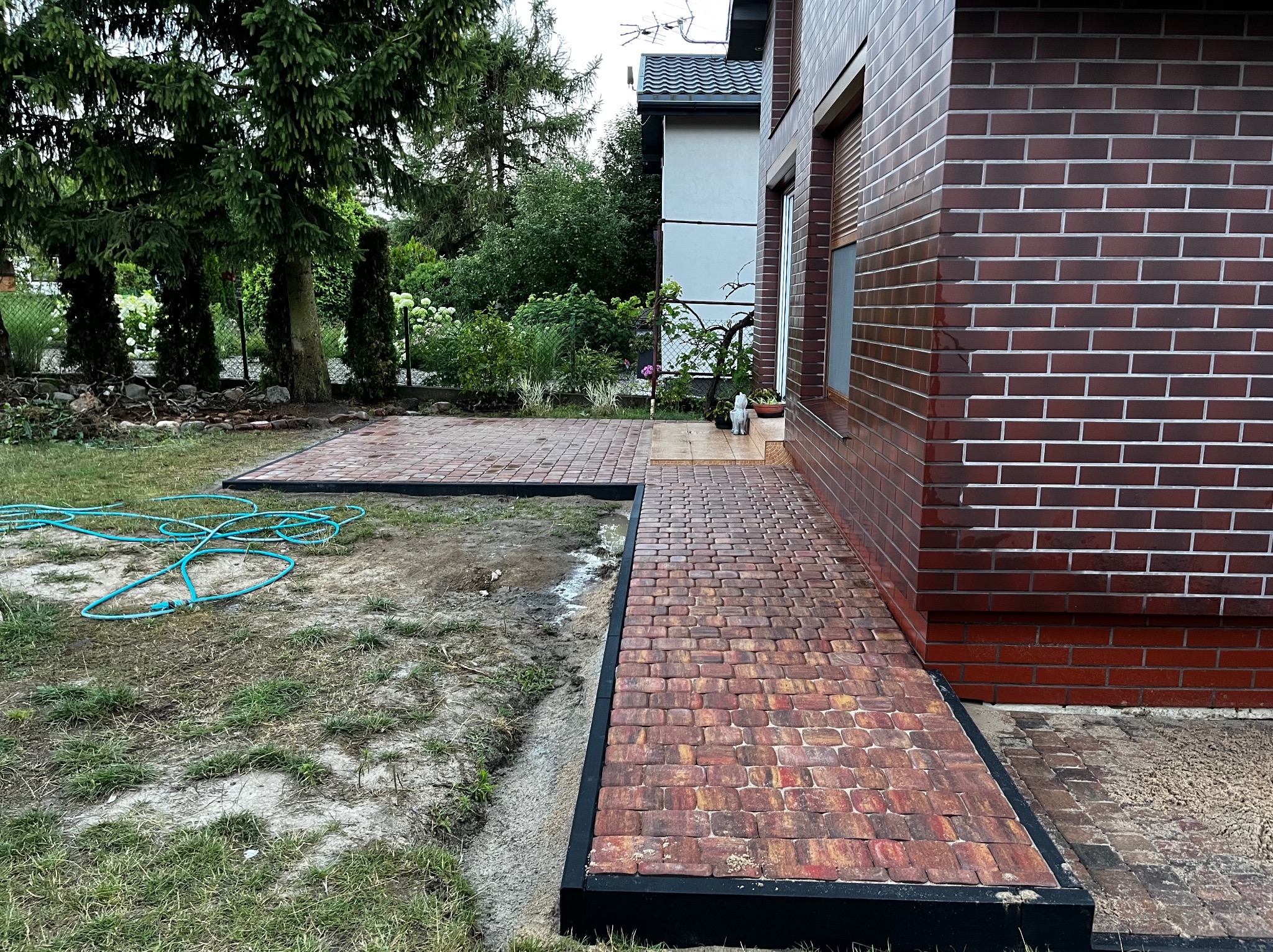 Czerwono-brązowa kostka brukowa tworząca ścieżkę i taras przy budynku z cegły, widoczny zwój niebieskiego węża ogrodowego na trawniku.