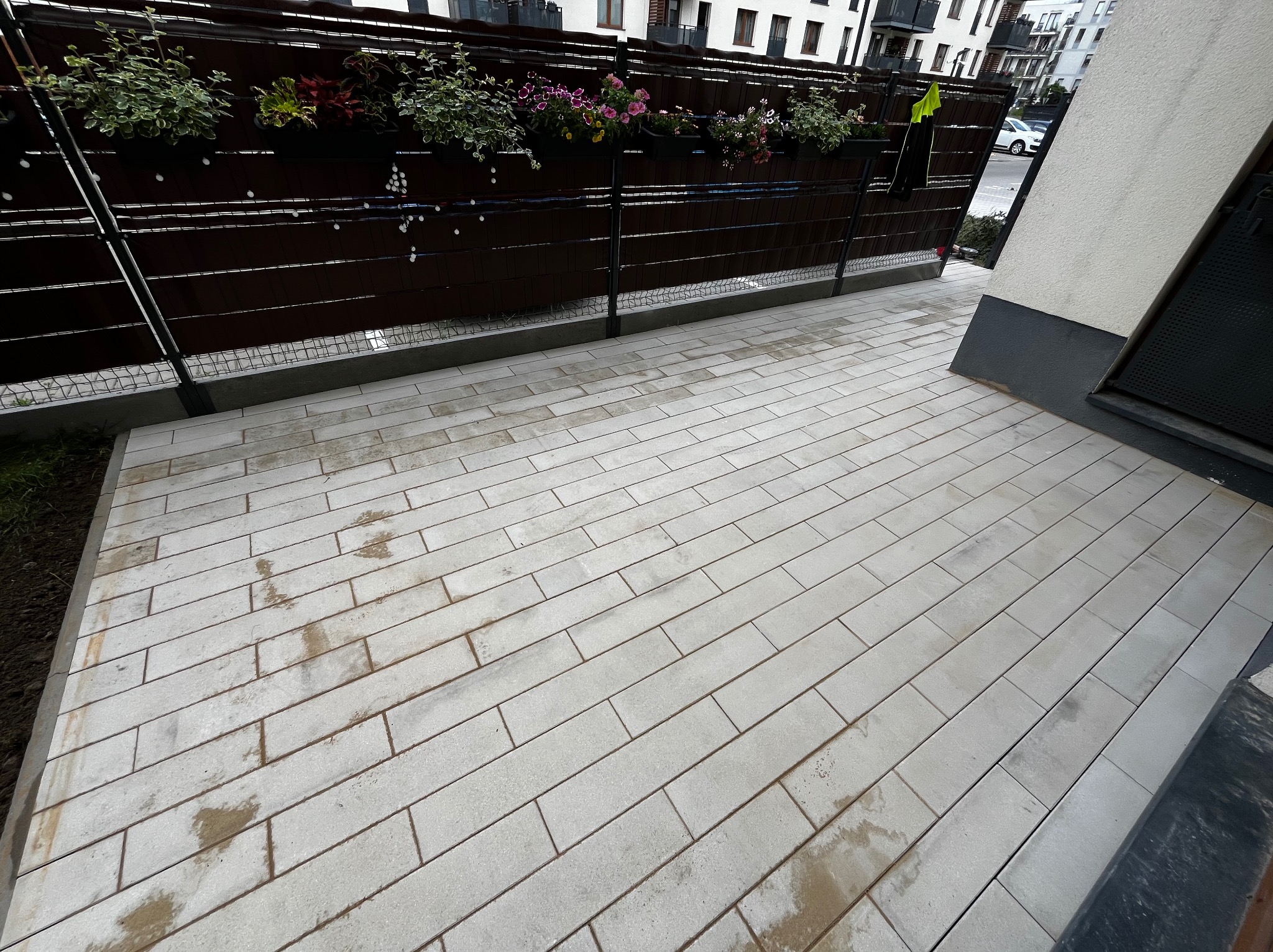 Świeżo ułożona, jasnoszara kostka brukowa w kształcie prostokątów, z widocznymi śladami wilgoci, przylegająca do ściany budynku i ogrodzenia z donicami z kwiatami.