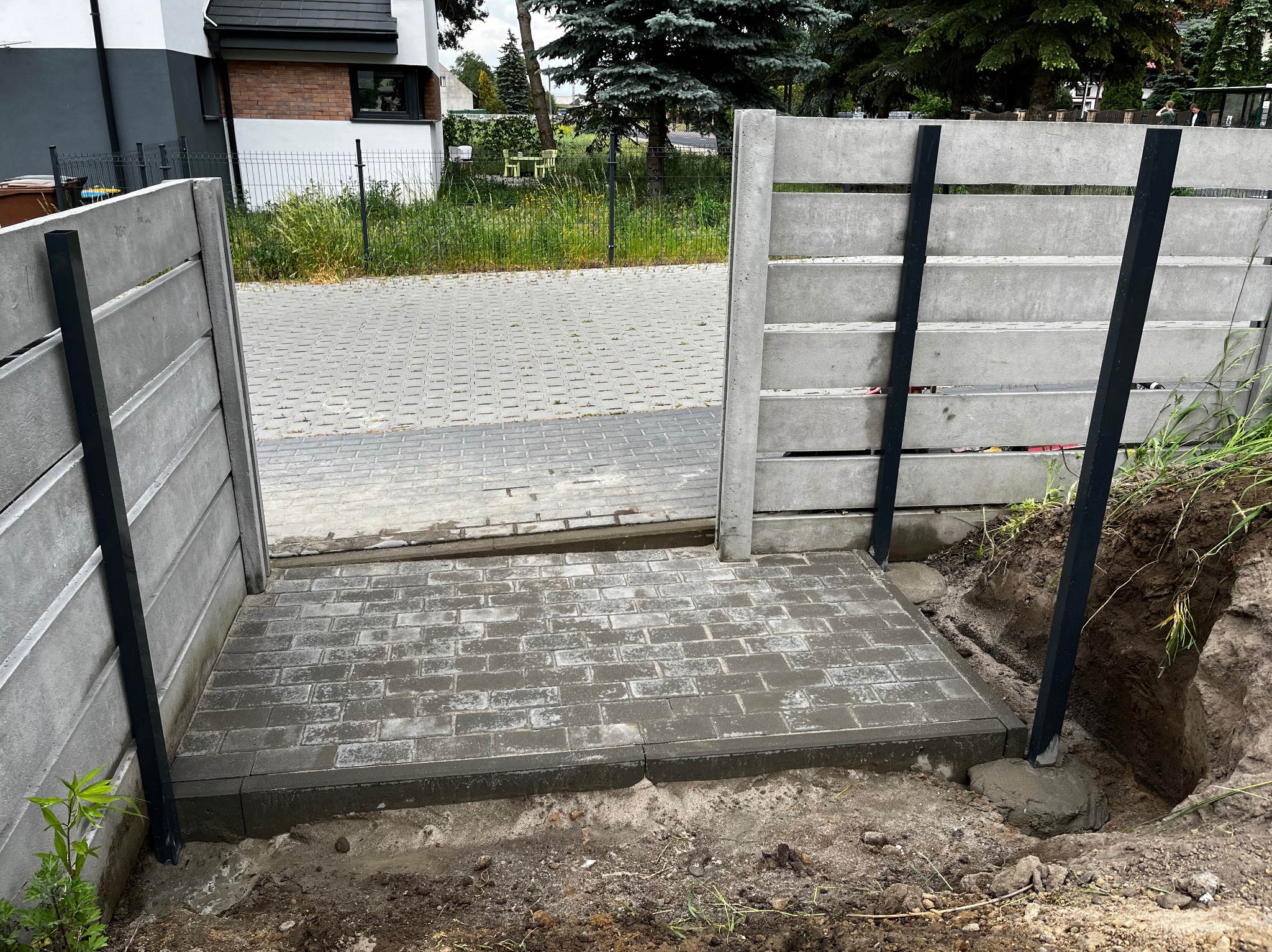 Fragment betonowego ogrodzenia panelowego z czarnymi słupkami i świeżo ułożona, wilgotna kostka brukowa prowadząca do furtki, widoczne wykopane dołki pod słupki.