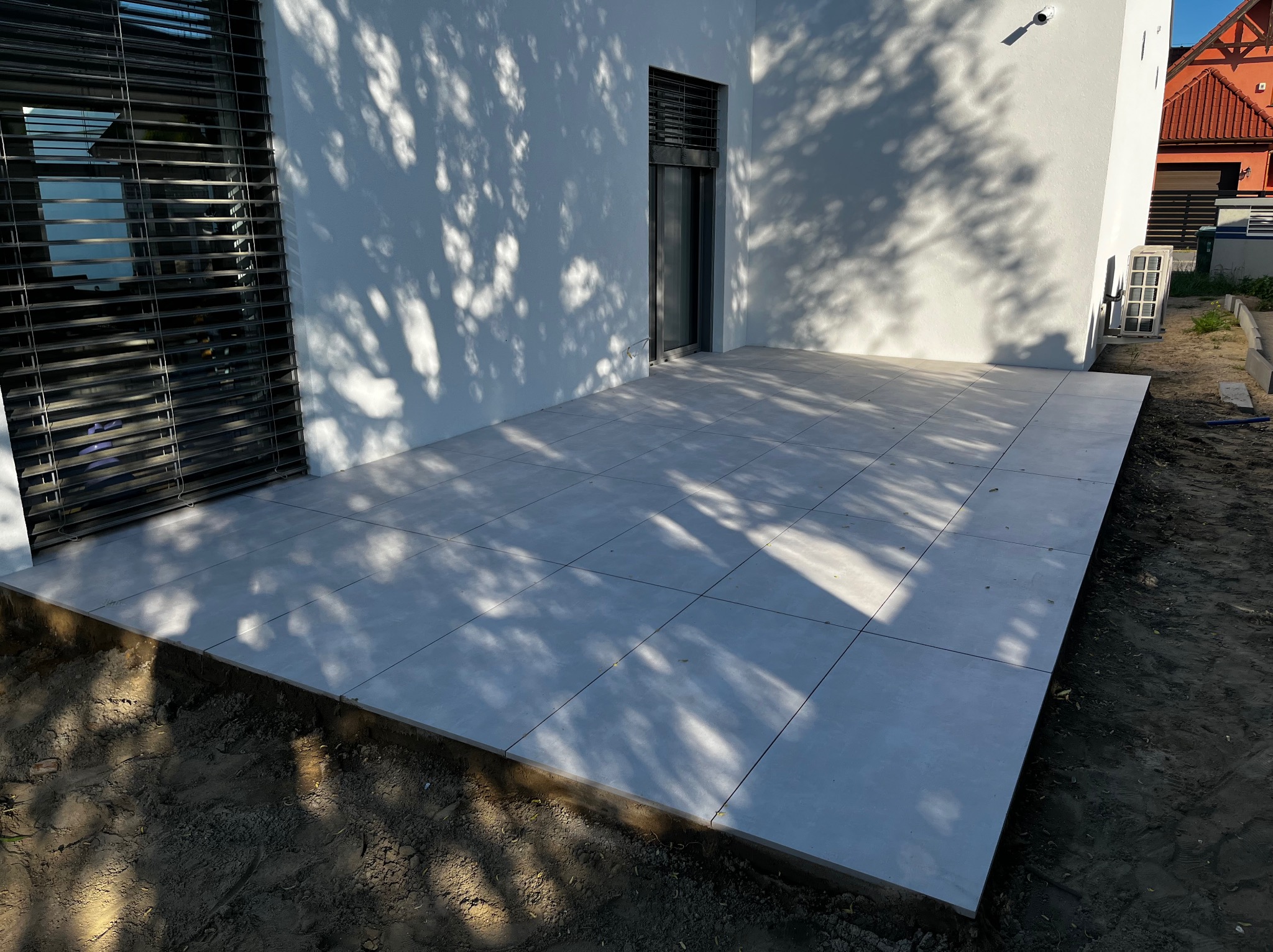 Jasnoszare płytki tarasowe ułożone przy białym budynku, z widocznymi cieniami drzew i ziemią wzdłuż krawędzi tarasu.