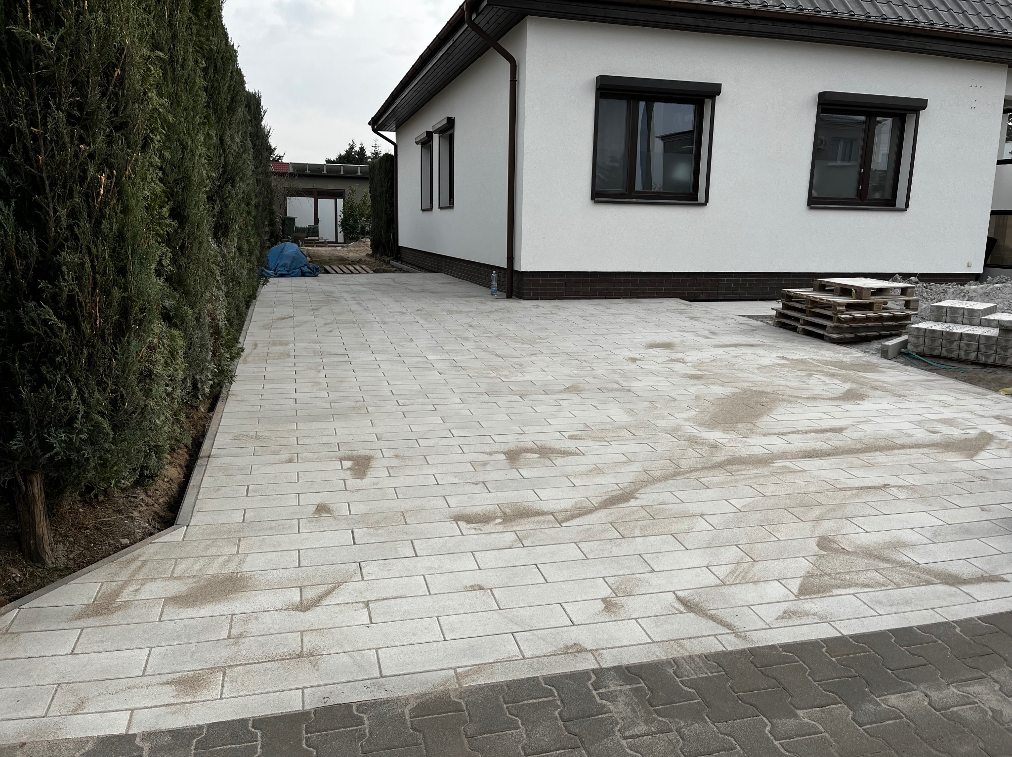 Ułożona jasnoszara kostka brukowa na podjeździe przed nowoczesnym domem z ciemnymi oknami, widoczne palety z kostką i wysoki żywopłot z boku.