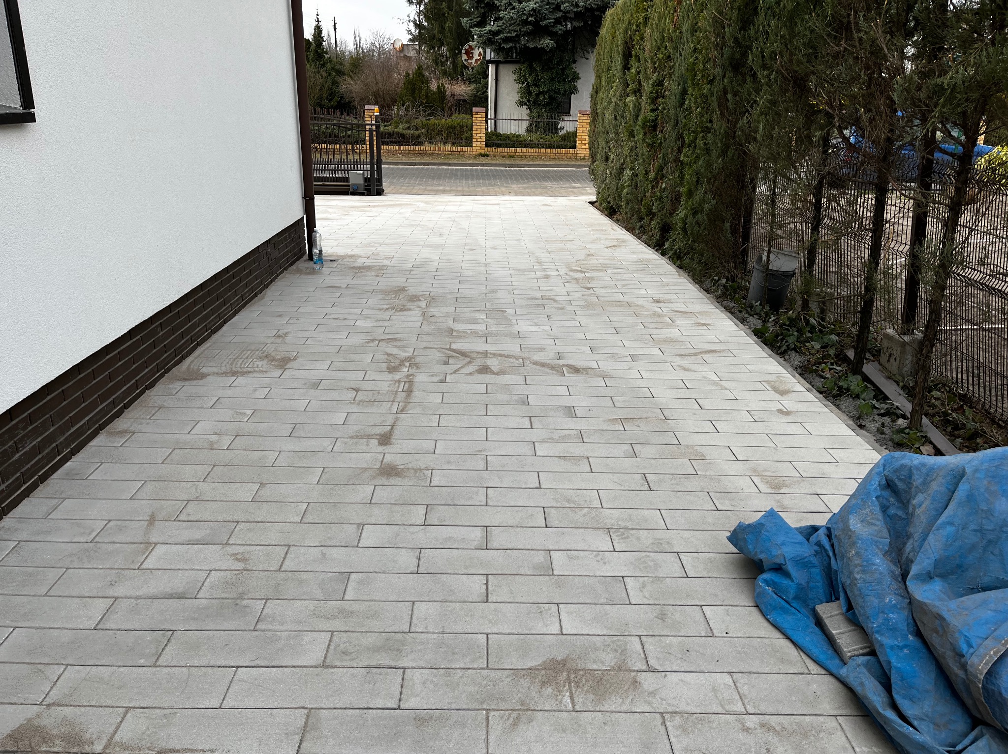 Świeżo ułożona, jasnoszara kostka brukowa w Poznaniu prowadząca od bramy wjazdowej do budynku, zabezpieczona częściowo niebieską plandeką. Widoczne ślady po pracy, sugerujące końcowy etap układania.